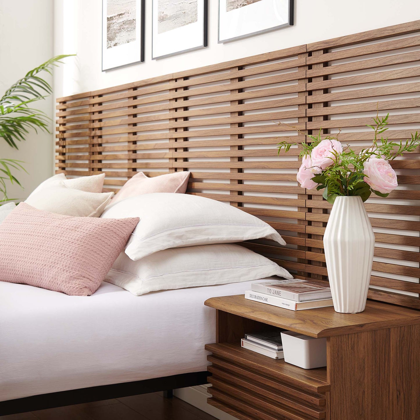 Walnut slatted wood wall-mounted king headboard with white bedding and a pink throw pillow; wooden nightstands with a vase of pink flowers and books.