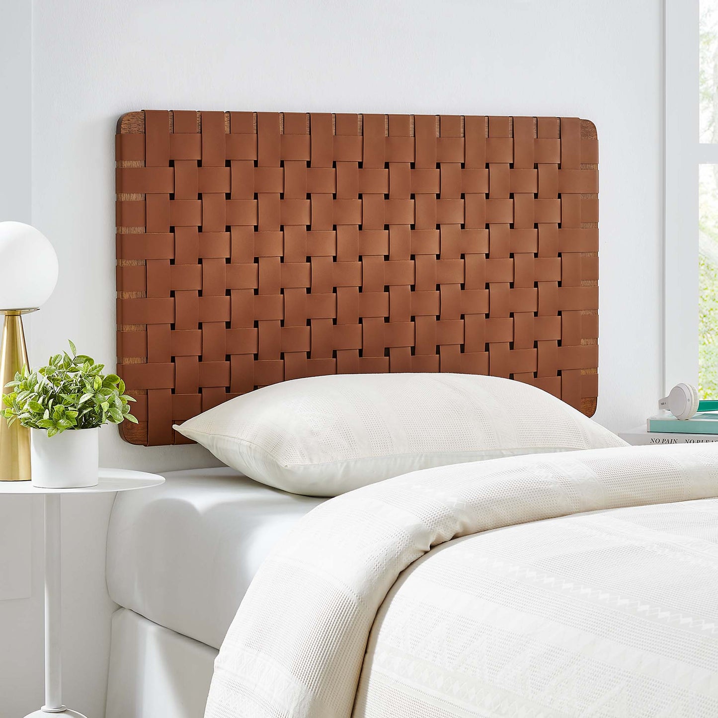 Walnut-brown vegan leather wall-mounted headboard on a white bed with cream bedding; white round nightstand, plant, and lamp beside.