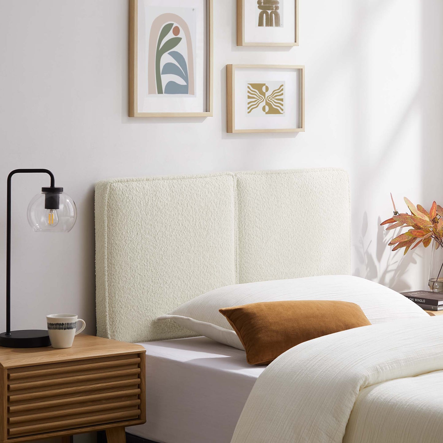 Cream boucle headboard with white bedding, brown throw pillow, wooden nightstand, black lamp, framed art.