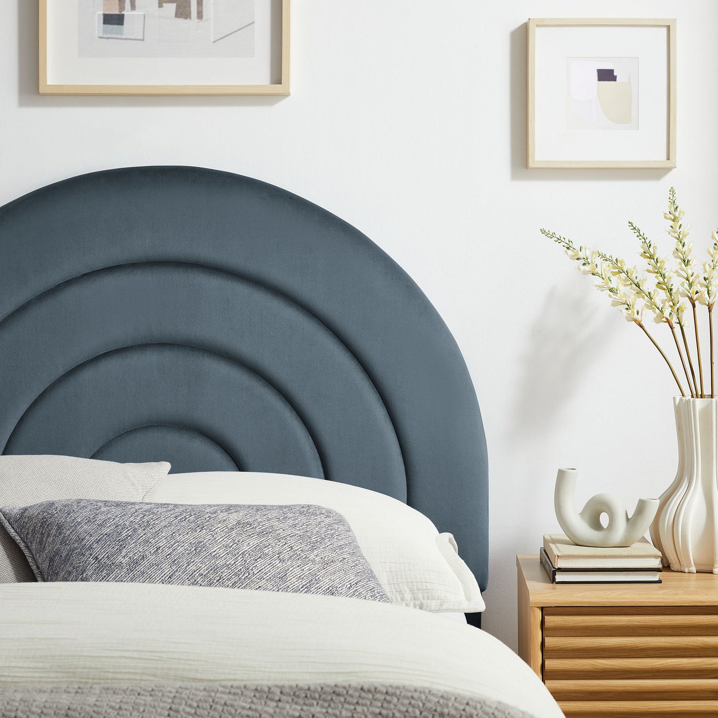 Circular navy upholstered headboard with concentric arches; white bedding, gray throw, wooden nightstand with vase of dried flowers.