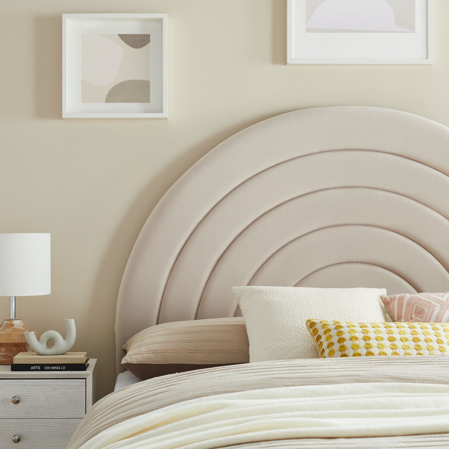 Oatmeal padded velvet arched headboard with concentric curves; beige bedding, mustard-yellow patterned pillow, and bedside lamp.