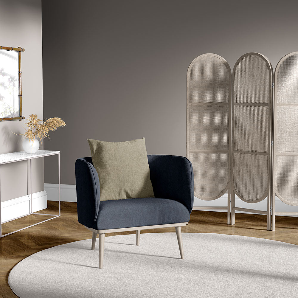 Navy blue curved armchair with light wood legs & beige pillow in a modern living room with a woven screen & cream rug.