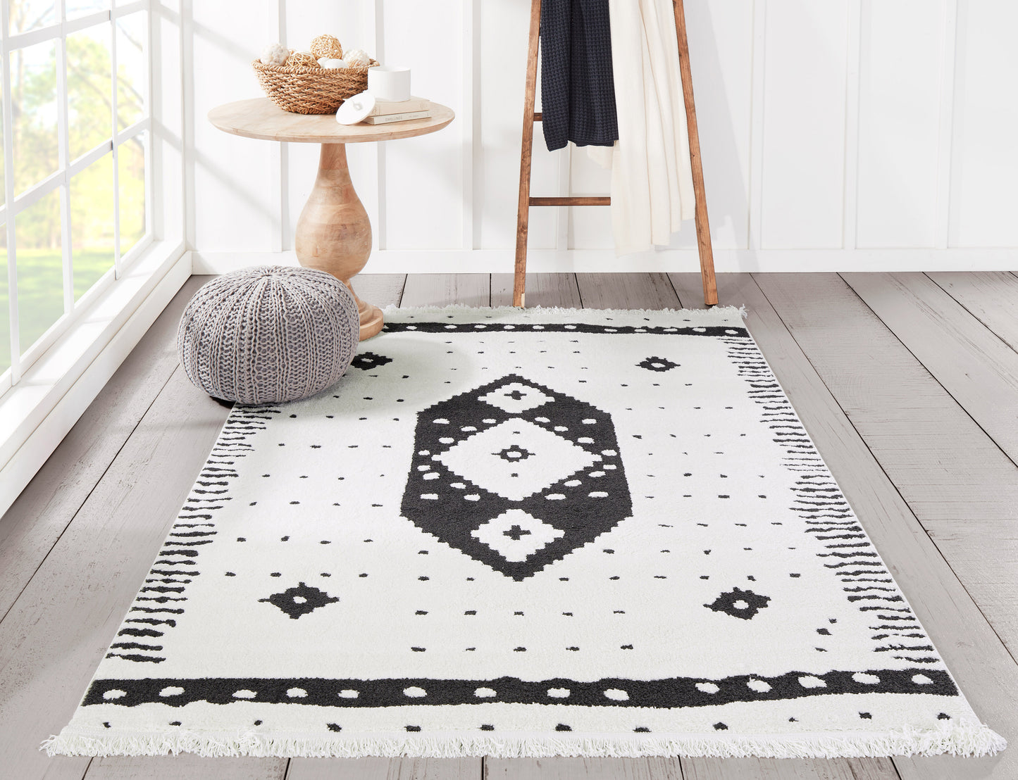 White rug with black geometric diamond motif and fringed edges in a bright room with a wooden table and knit pouf.