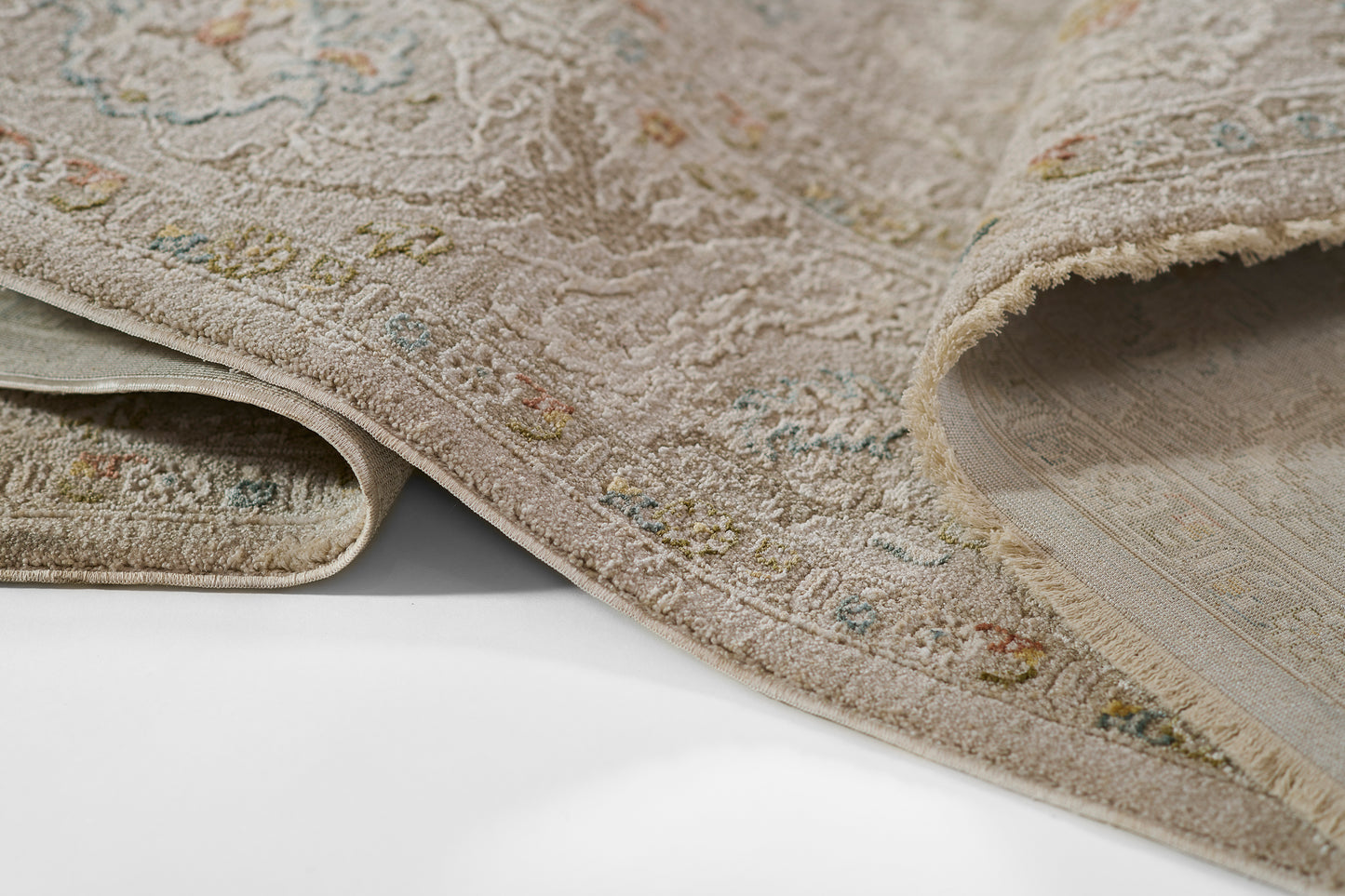 Beige ornate rug with subtle multicolor floral motifs, plush pile, folded edge showing backing.
