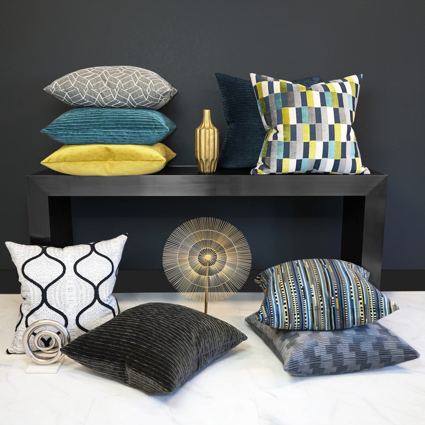 Mid-mod luxe: dark wall, black console with stacked teal, gray, and yellow pillows; gold vase; sunburst decor; blue and gray floor cushions.