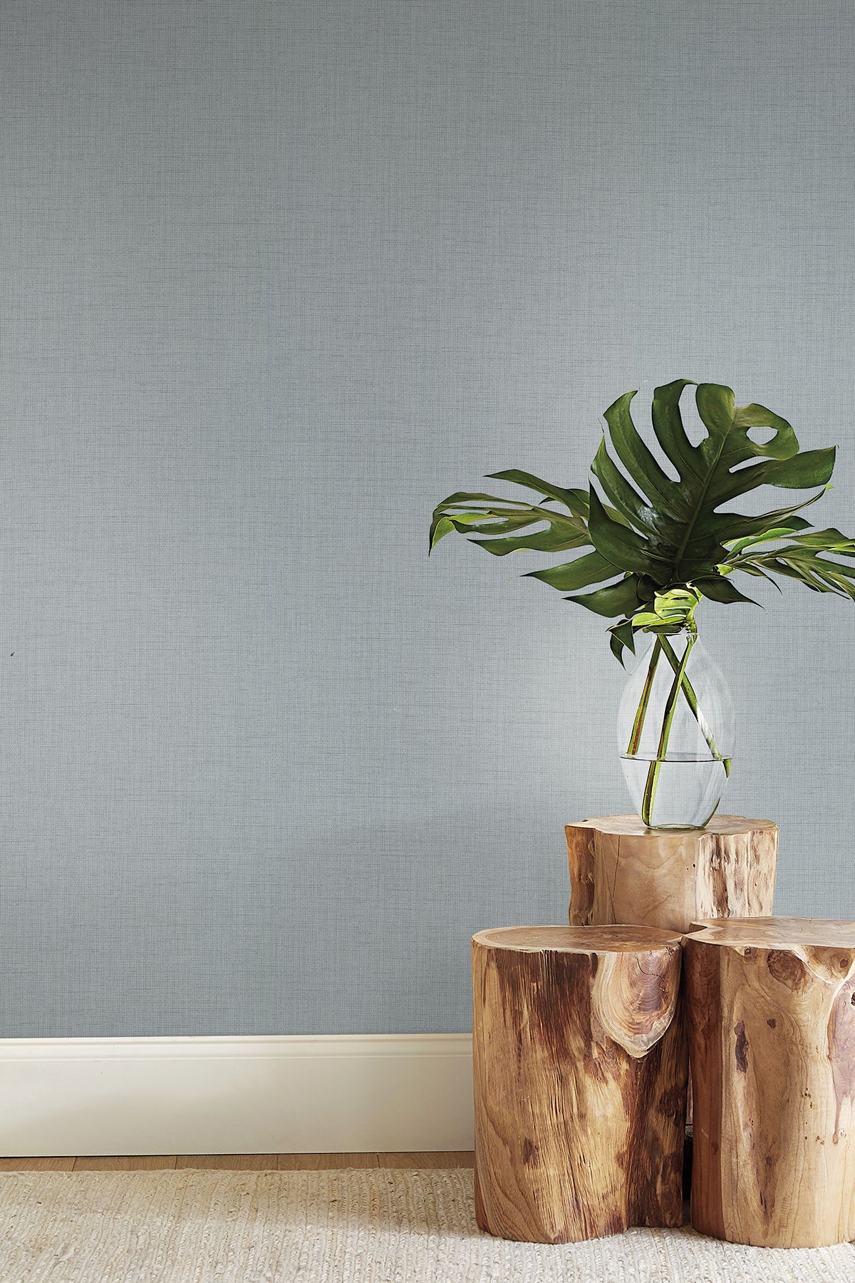 Clear glass vase with water and monstera leaves atop stacked natural wood stumps against a gray textured wall.