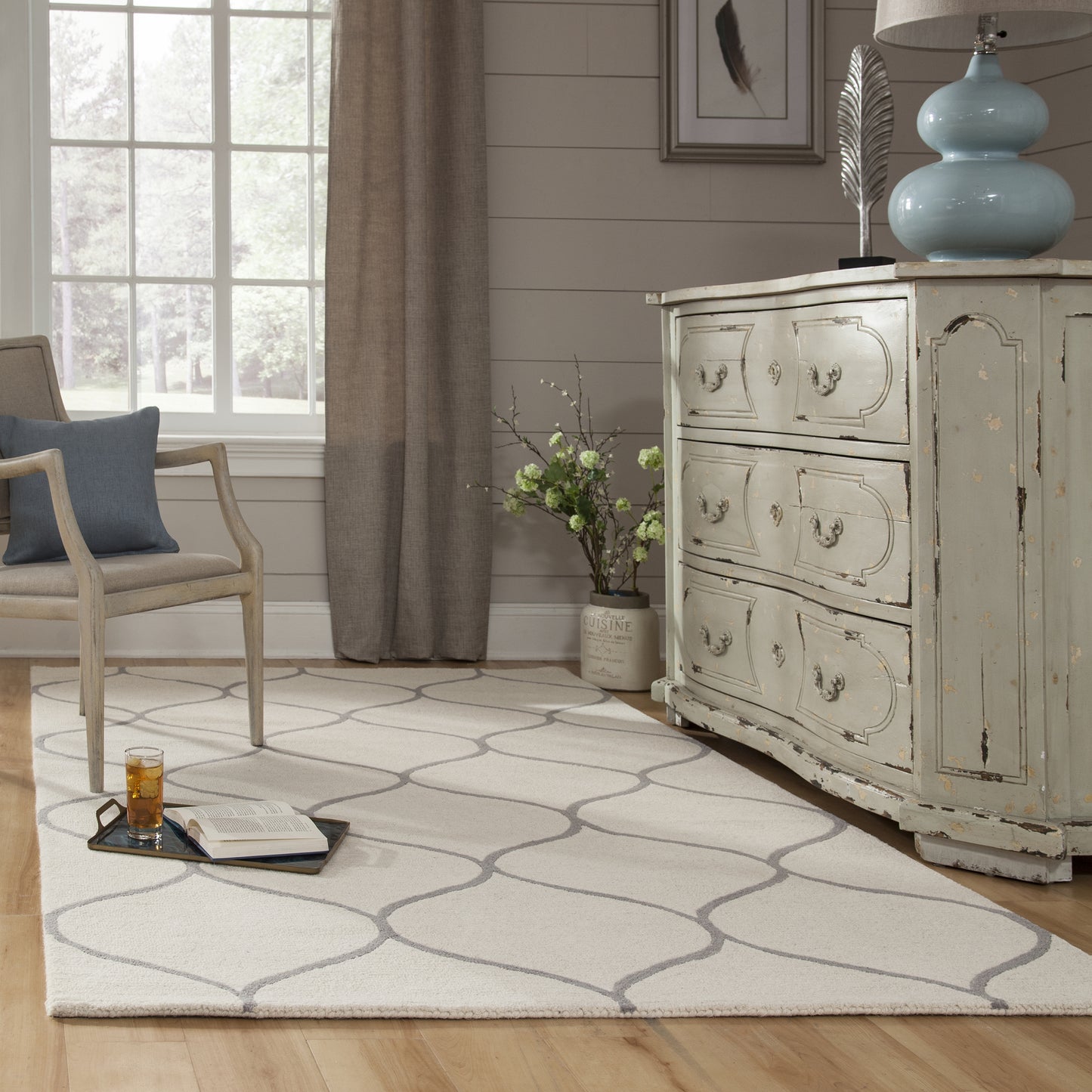 Shabby chic living room with a distressed gray dresser, pale blue lamp, beige armchair, and geometric lattice rug.