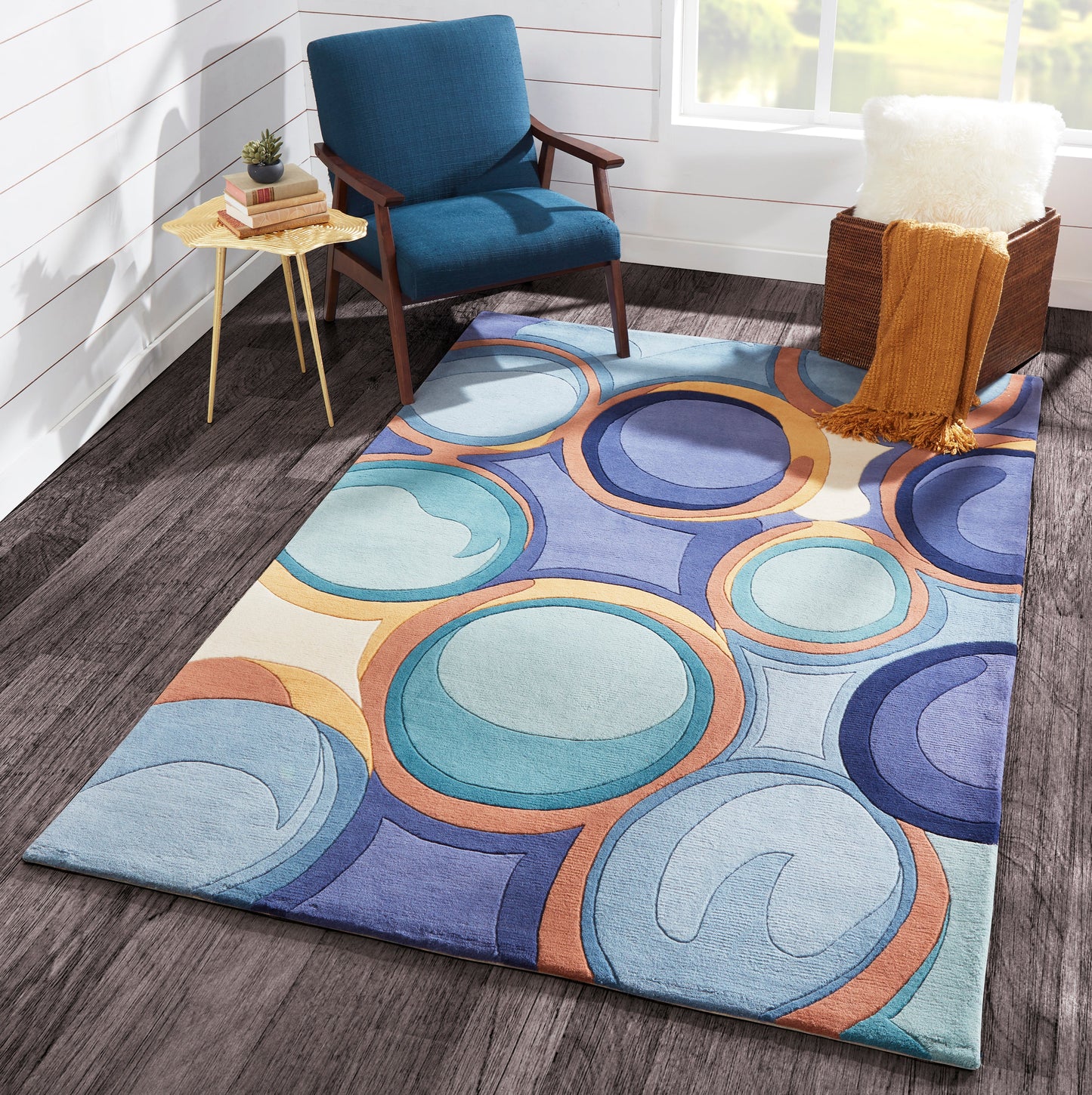 Colorful circular-pattern area rug in blues, teals and orange under a blue upholstered lounge chair with wooden arms.