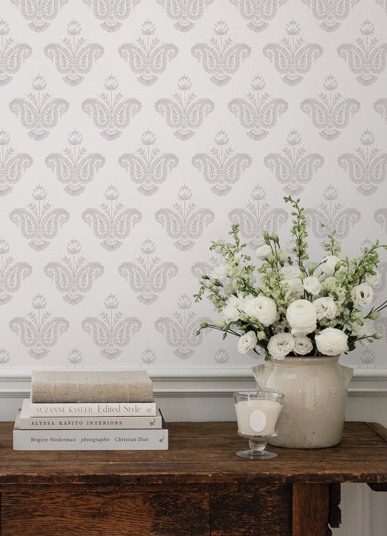 Soft gray botanical linen wallpaper, rustic wooden console, stack of cream books, white floral ceramic vase, and a candle.