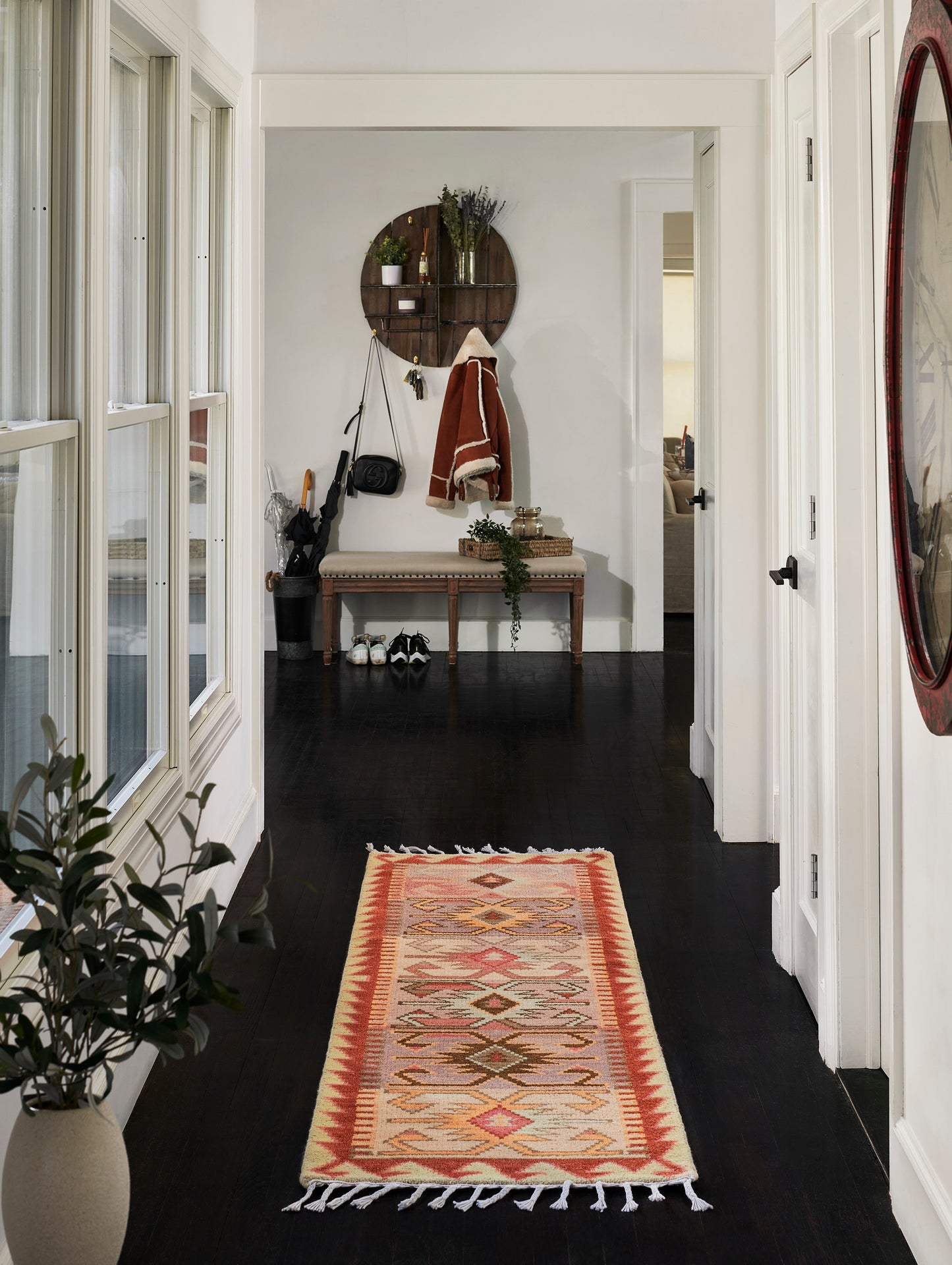 Geometric woven area rug in coral, cream and orange with fringed ends, down a dark-wood hallway beside a bench.