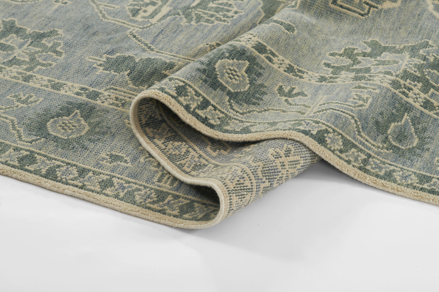 Blue-green oriental rug with cream fringe, intricate floral border; rolled corner shows soft woven texture.