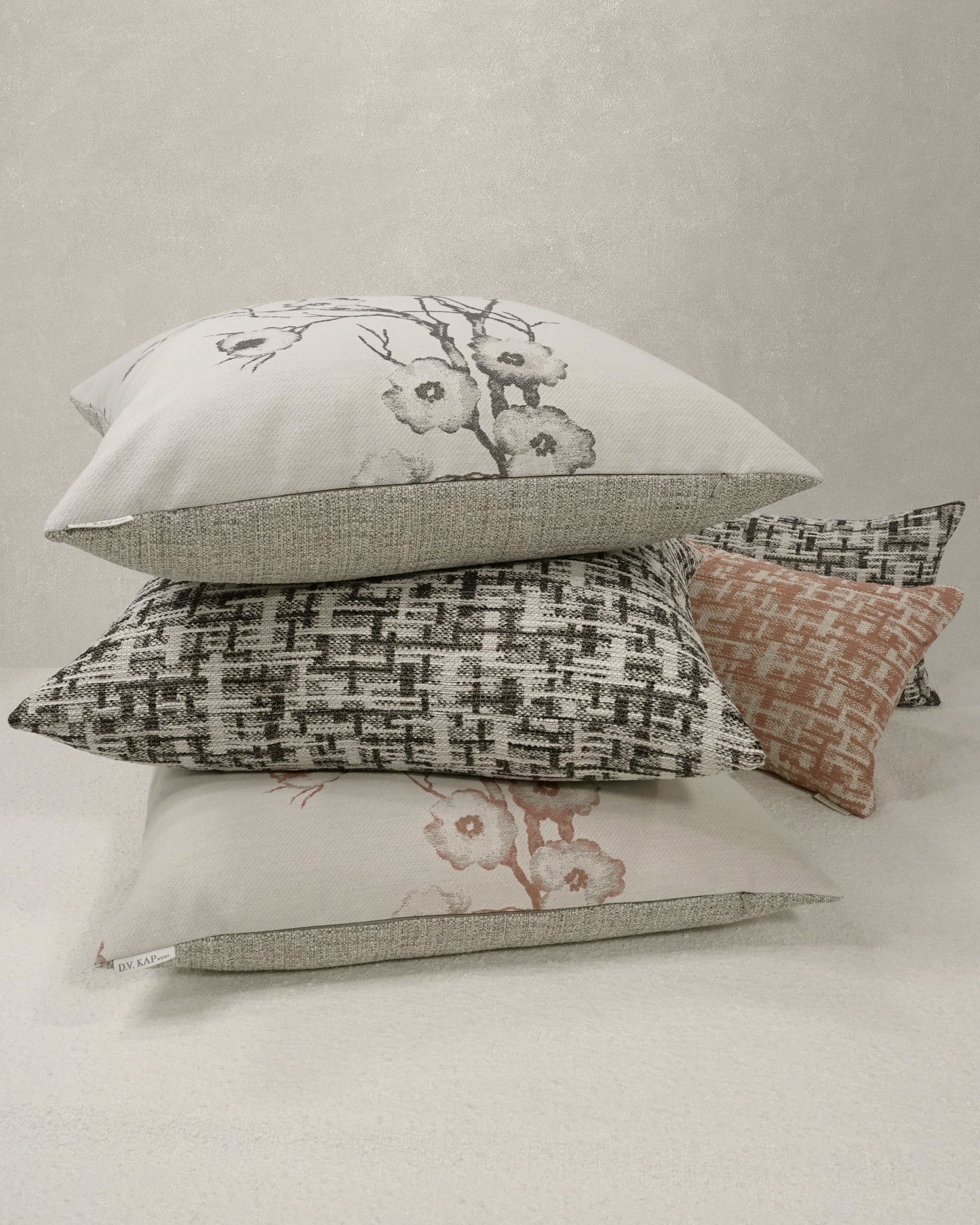 Stack of decorative rectangular cushions: ivory floral, beige textured, black & white woven, pale pink blossom, with small peach accent.