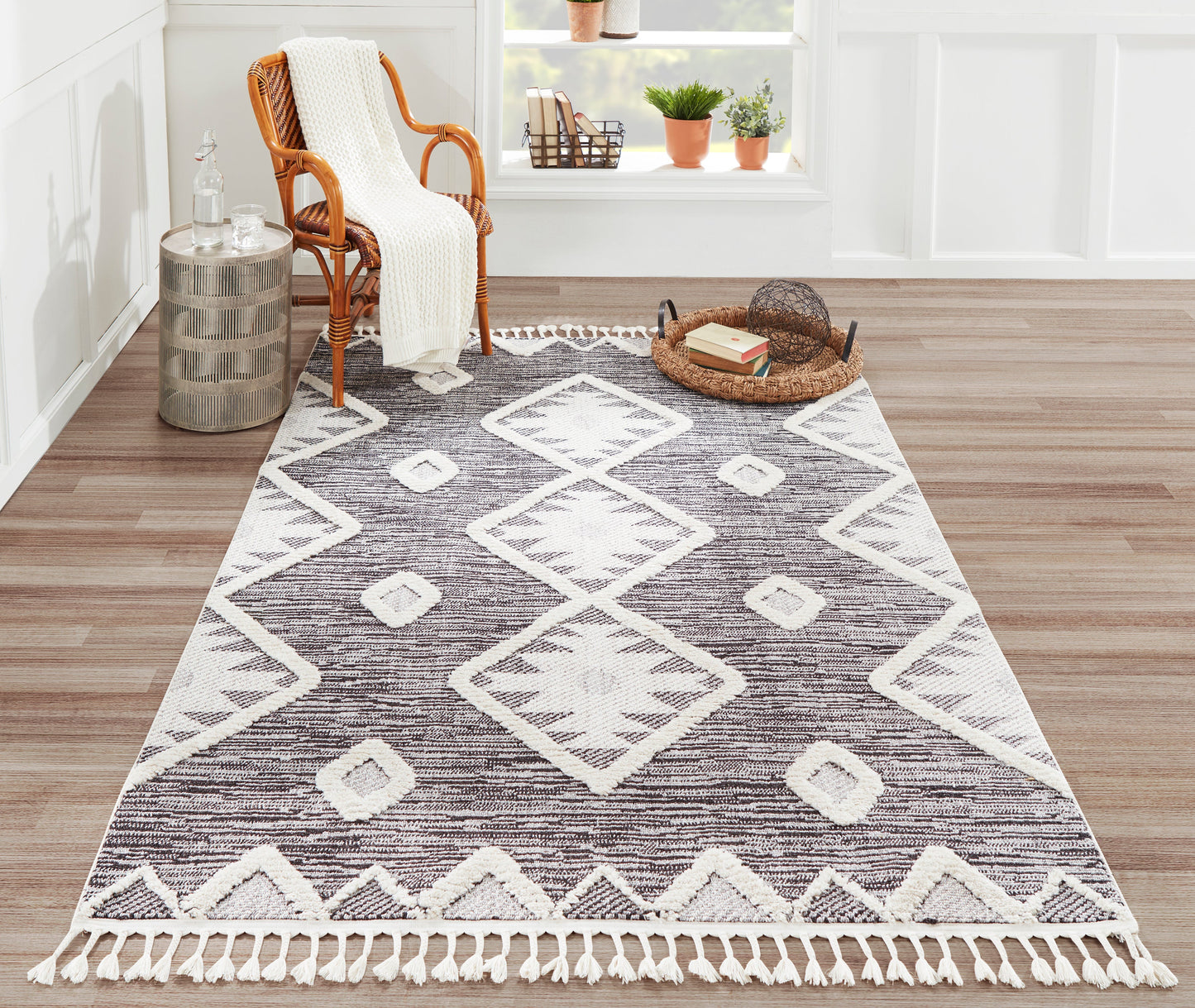 Geometric area rug with white diamond motifs and fringe, charcoal brown tones on a wood floor beside a wicker chair.