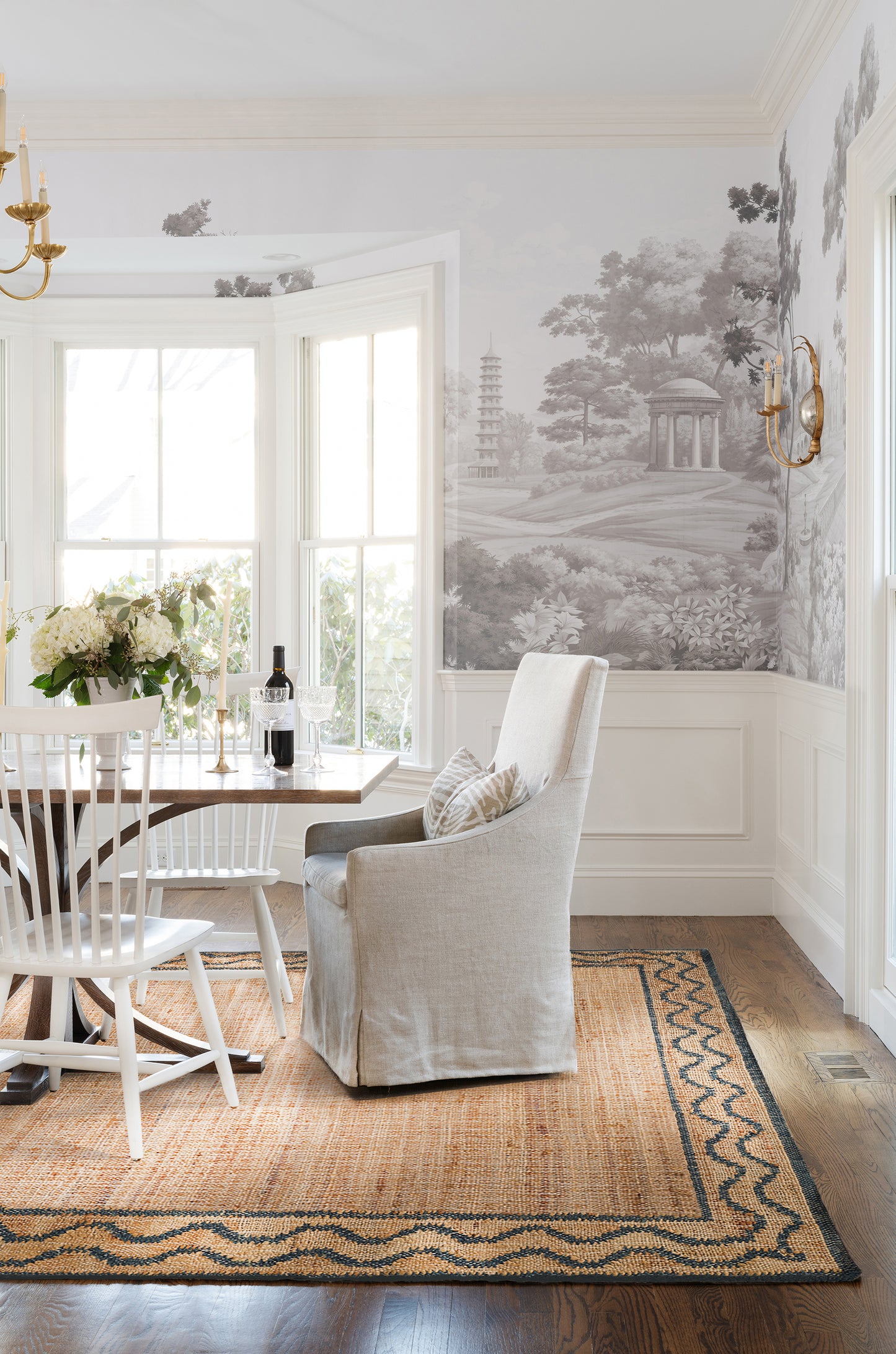 Beige slipcovered armchair beside wooden dining table; white Windsor chairs, landscape wallpaper, brass sconces, wine and flowers.
