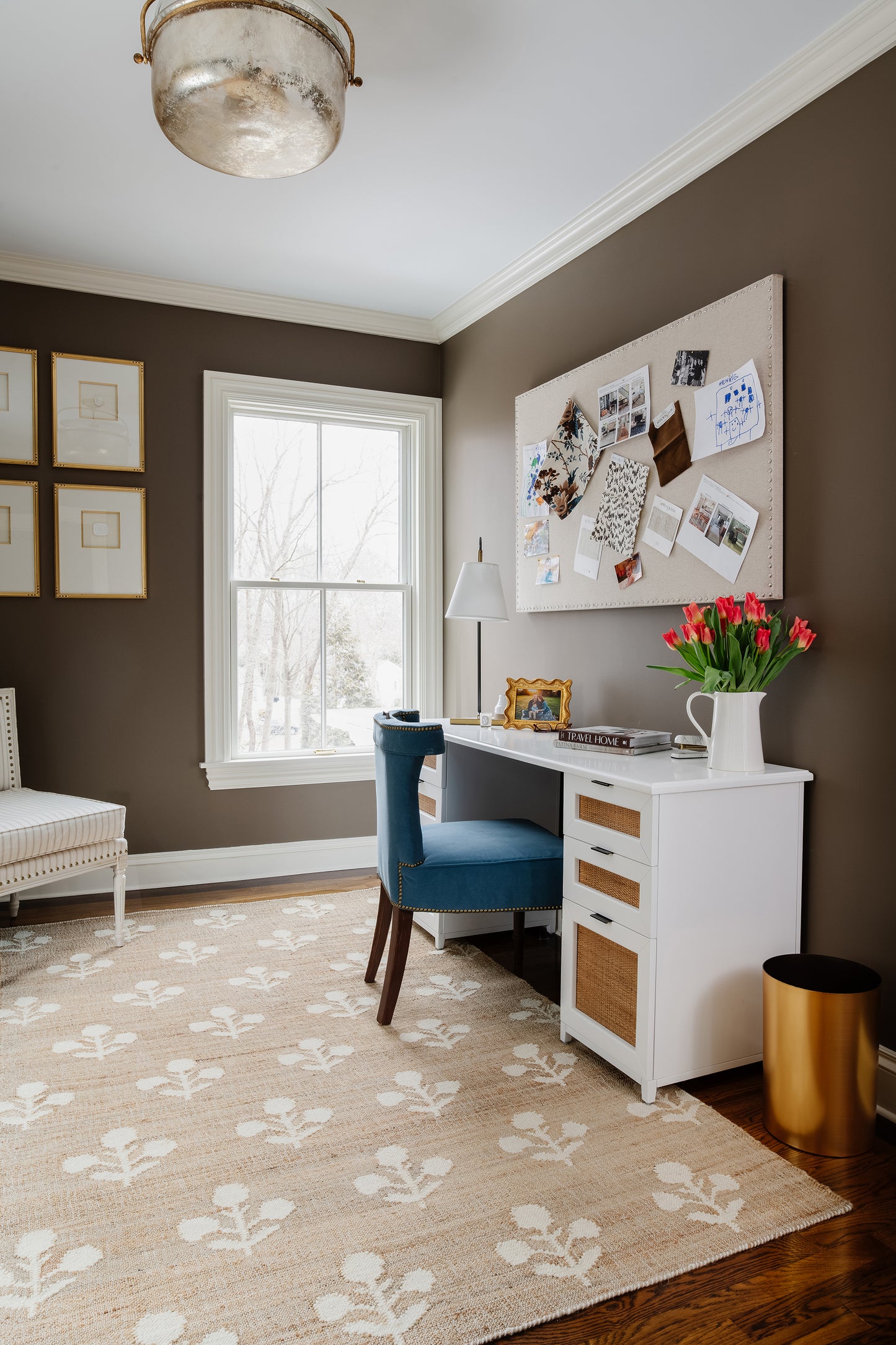 Brown-walled home office with white desk, blue velvet chair, corkboard, tulips in a pitcher, beige leaf rug.