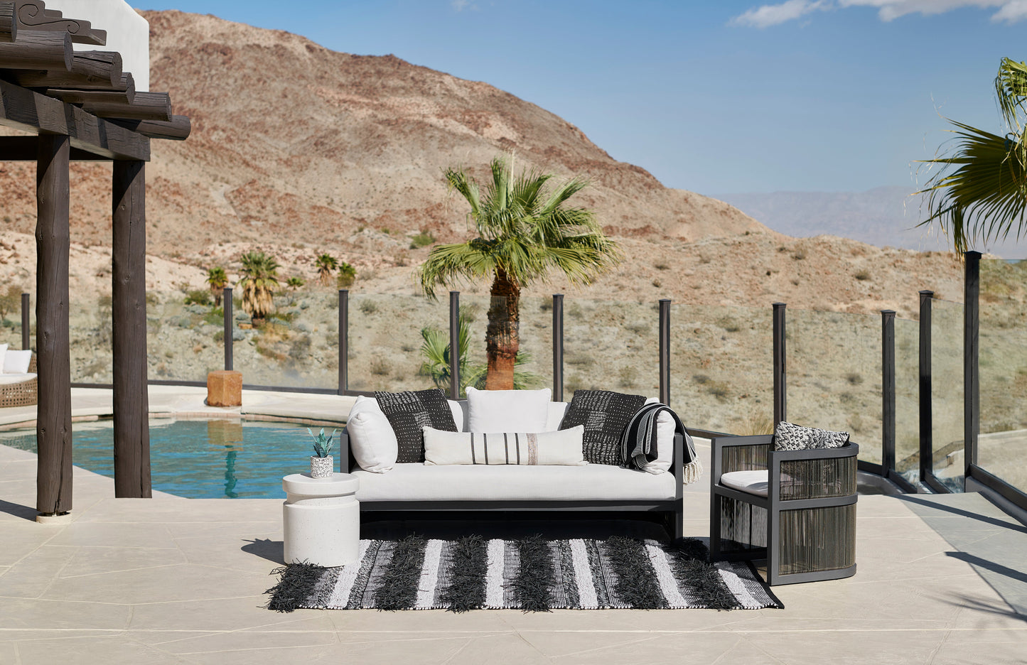 Outdoor modern patio with white cushioned sectional, black wicker chair, striped rug, pool and glass railing.