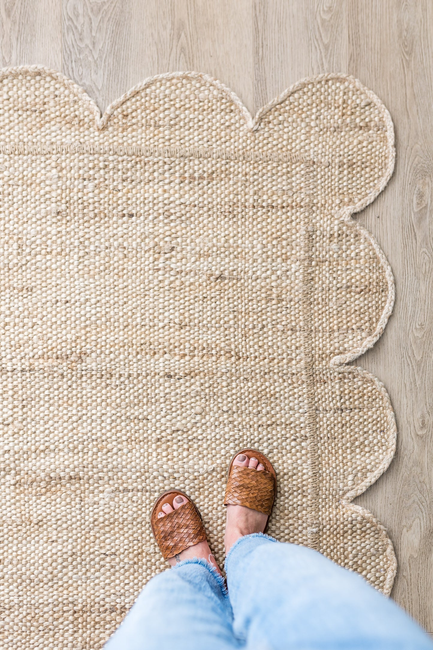 Natural jute rug with scalloped edge, top-down view; feet in brown woven sandals and light-wash jeans on a wooden floor.