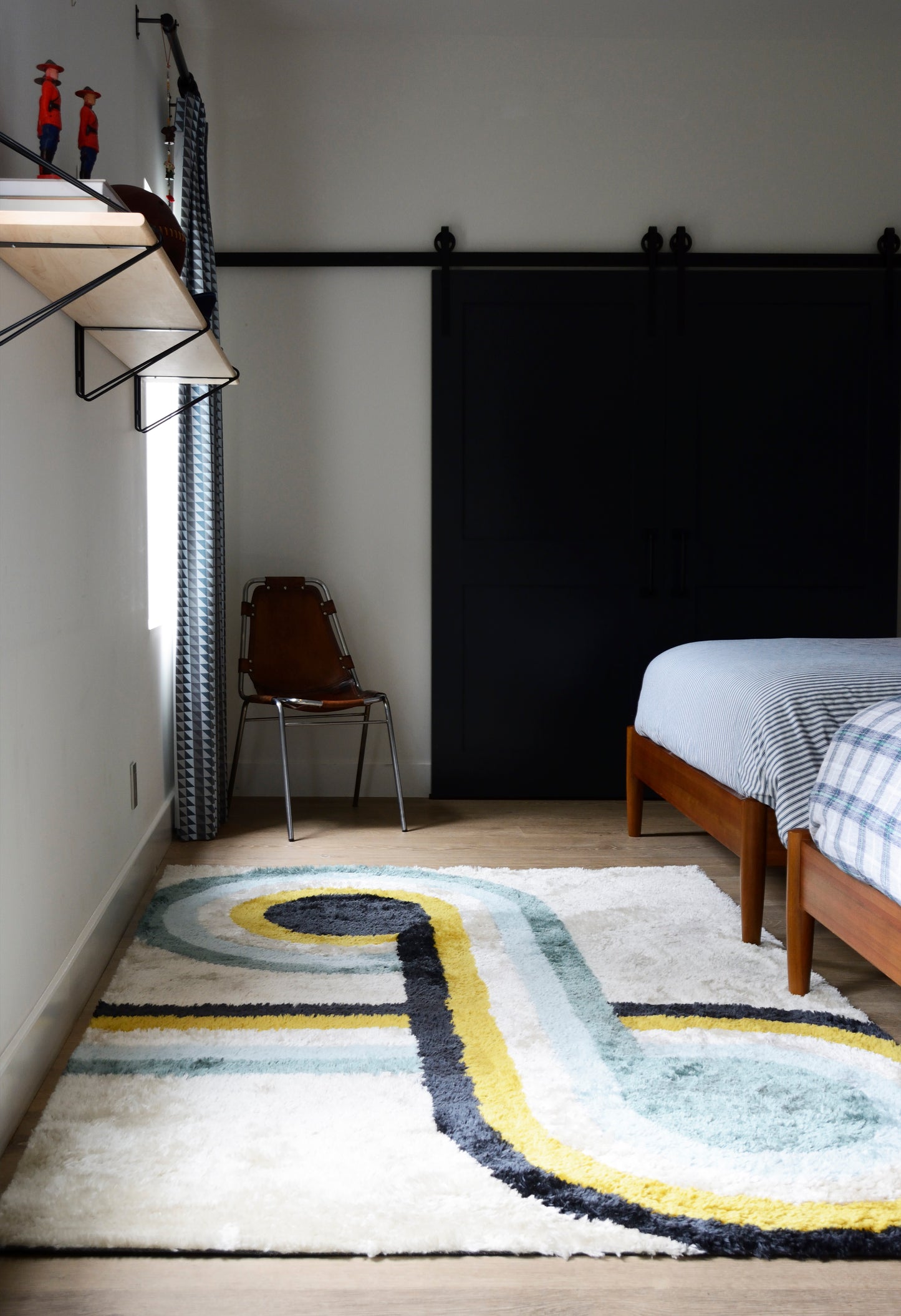 Cozy modern bedroom with two twin beds in wood frames, blue-check bedding, black sliding barn door, striped rug, metal chair.