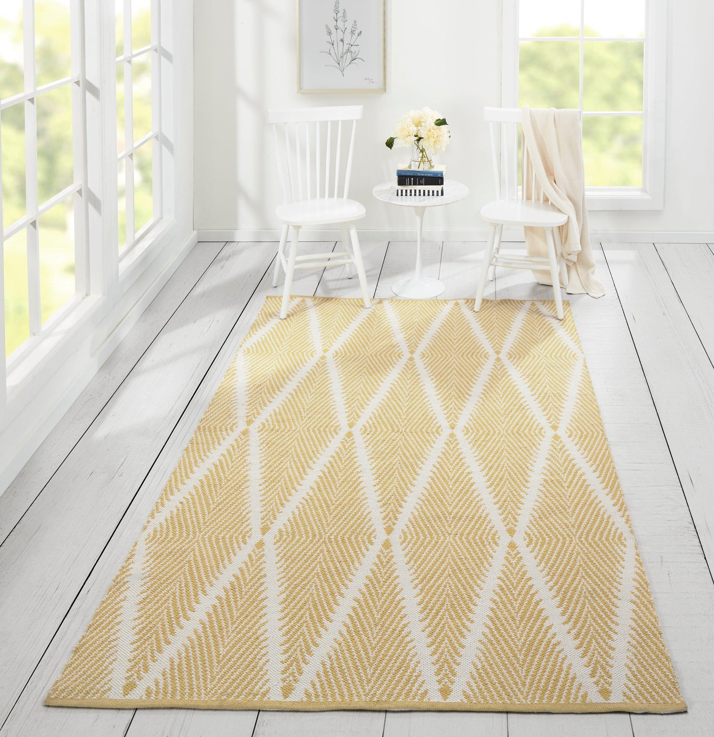 Bright sunlit white room with a yellow geometric area rug featuring white diamonds, two white chairs & a small table.