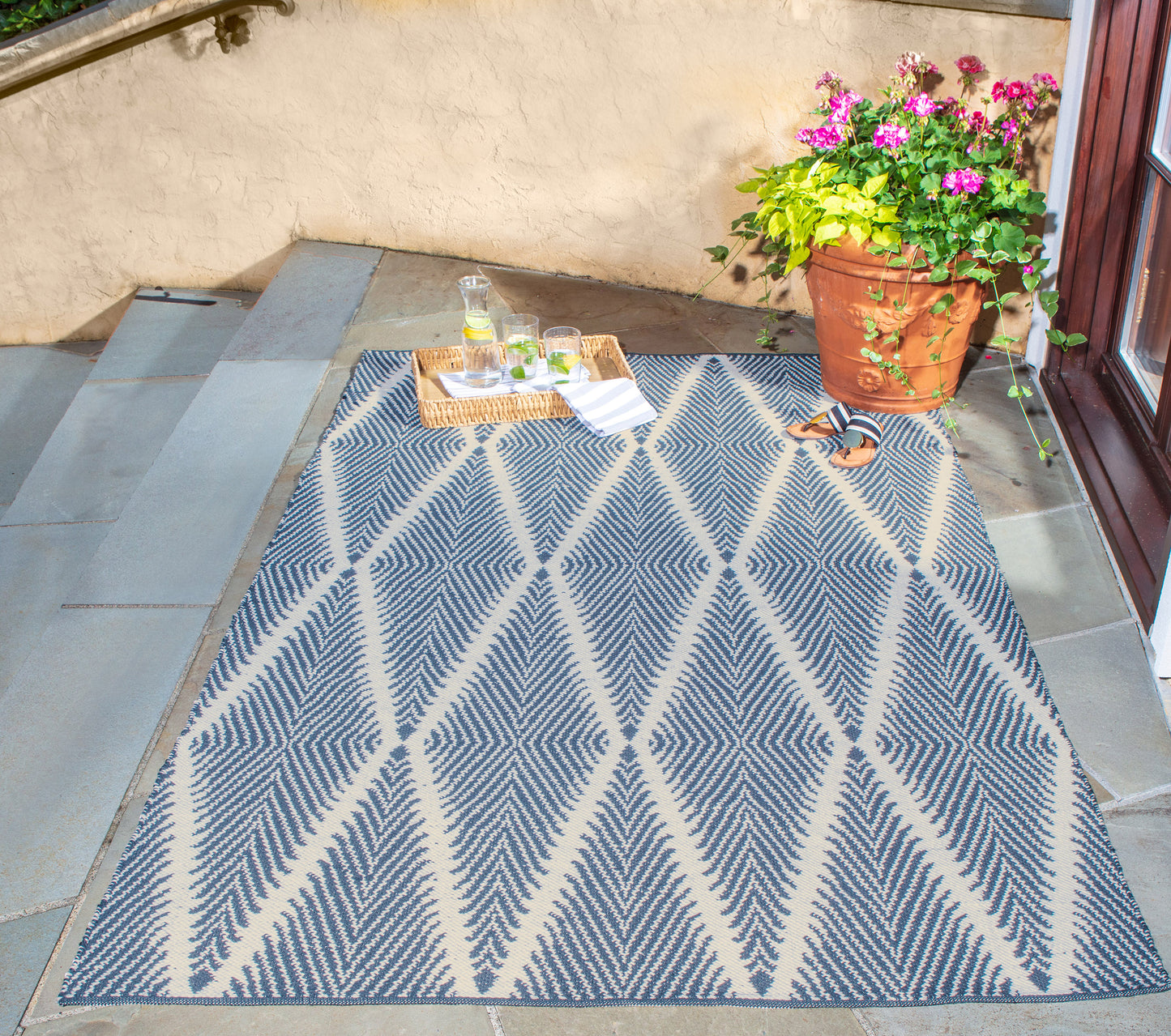 Blue and white geometric outdoor rug on stone steps by a door, with a tray of drinks & a potted pink-flowering plant.