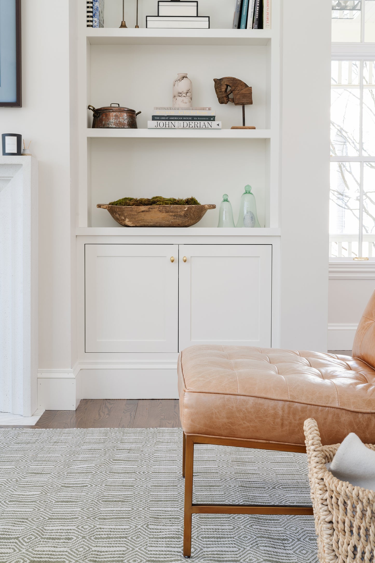 White built-in bookshelf with cabinet doors and decorative items; tan tufted leather ottoman in a bright living room.