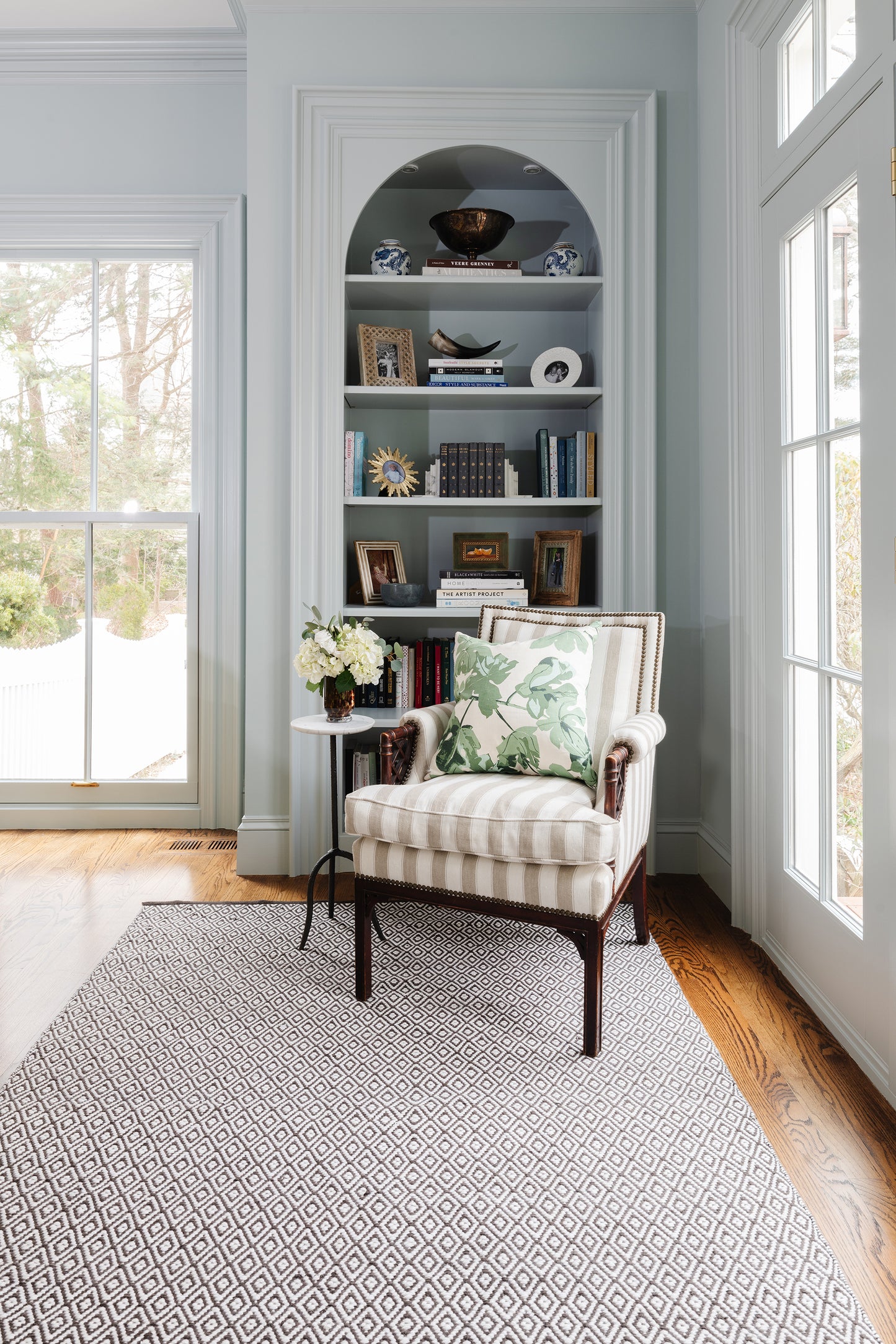 Cozy reading nook: striped beige armchair with floral pillow, light blue built-in arch bookshelf, side table with flowers, geometric rug.