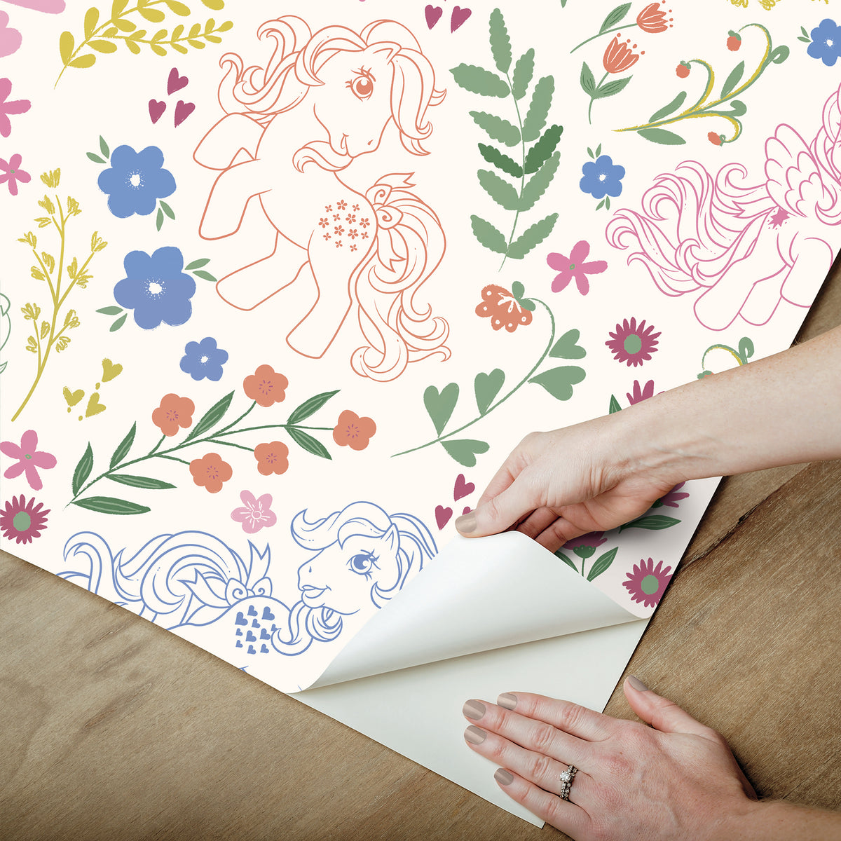 Colorful floral and pony-themed wallpaper sample being peeled at the corner, exposing white backing; hands hold the sheet.