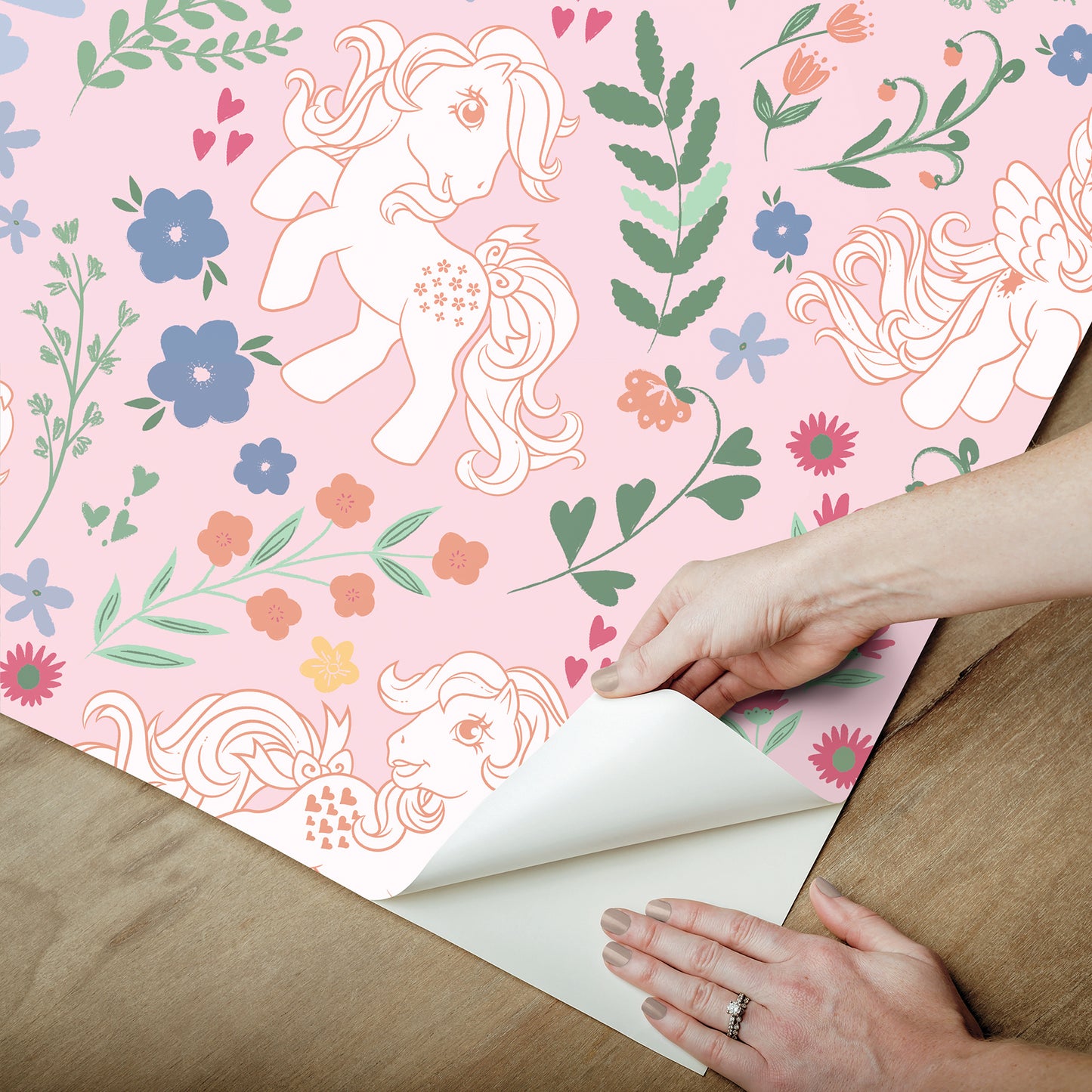 Pastel pink wallpaper with cartoon ponies and flowers; hands peel back a corner to reveal white backing.