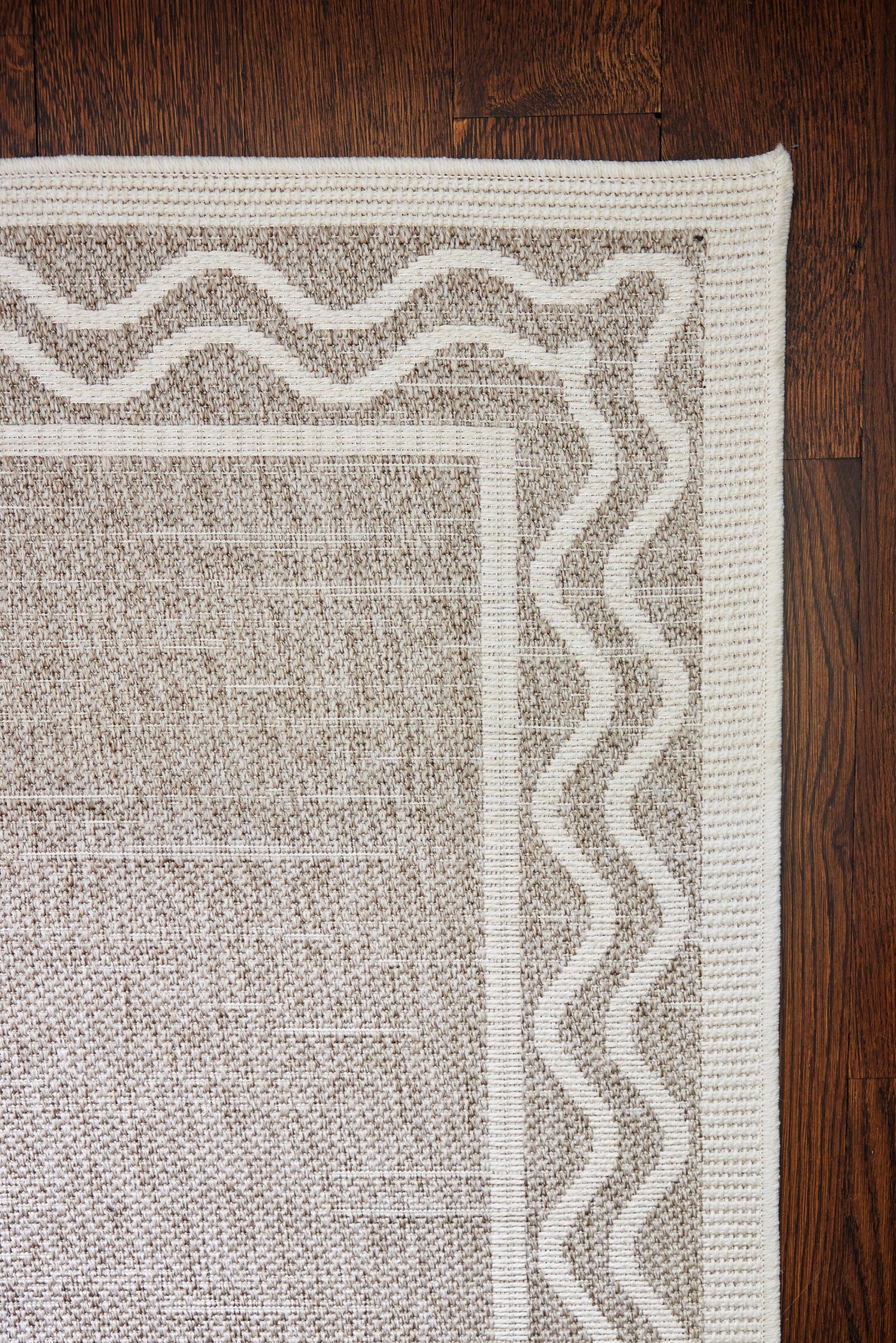 Neutral gray-beige flatweave rug with cream wavy border and textured central panel on dark wood floor.
