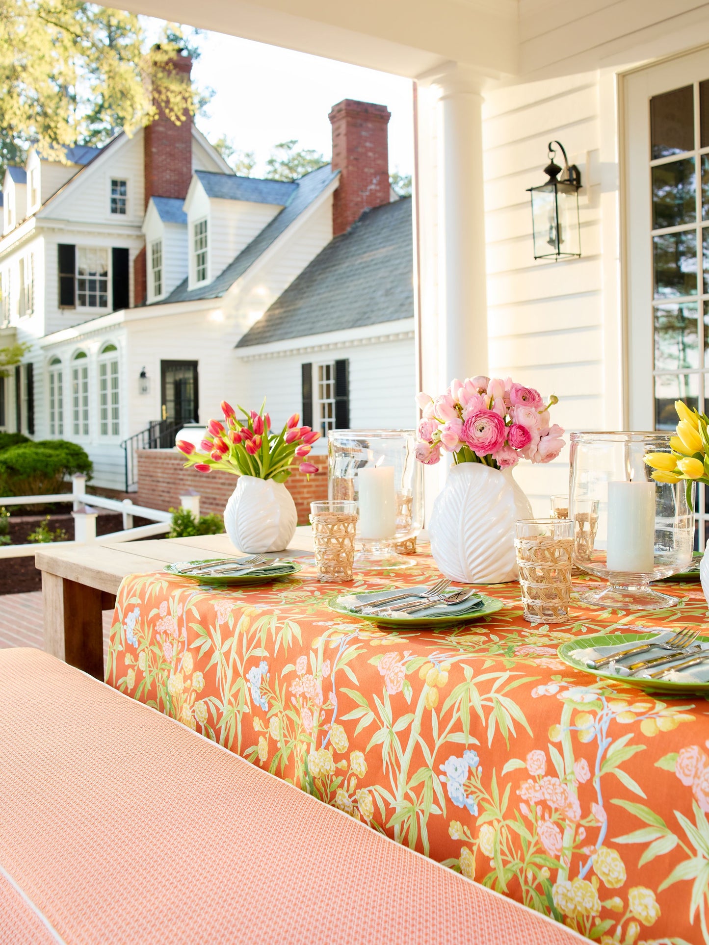 Outdoor porch dining with orange floral tablecloth, peach bench, white vases of pink flowers, green plates, candles.