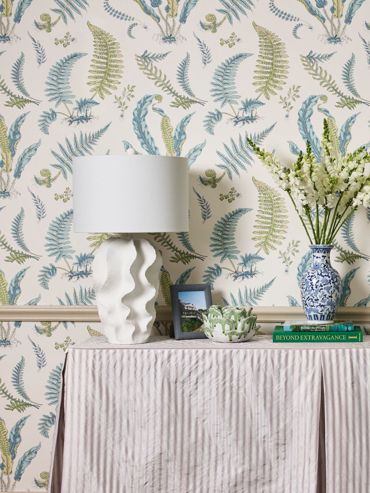 Pastel fern wallpaper, white sculptural lamp, pink striped table, blue-and-white vase with white flowers, green books.