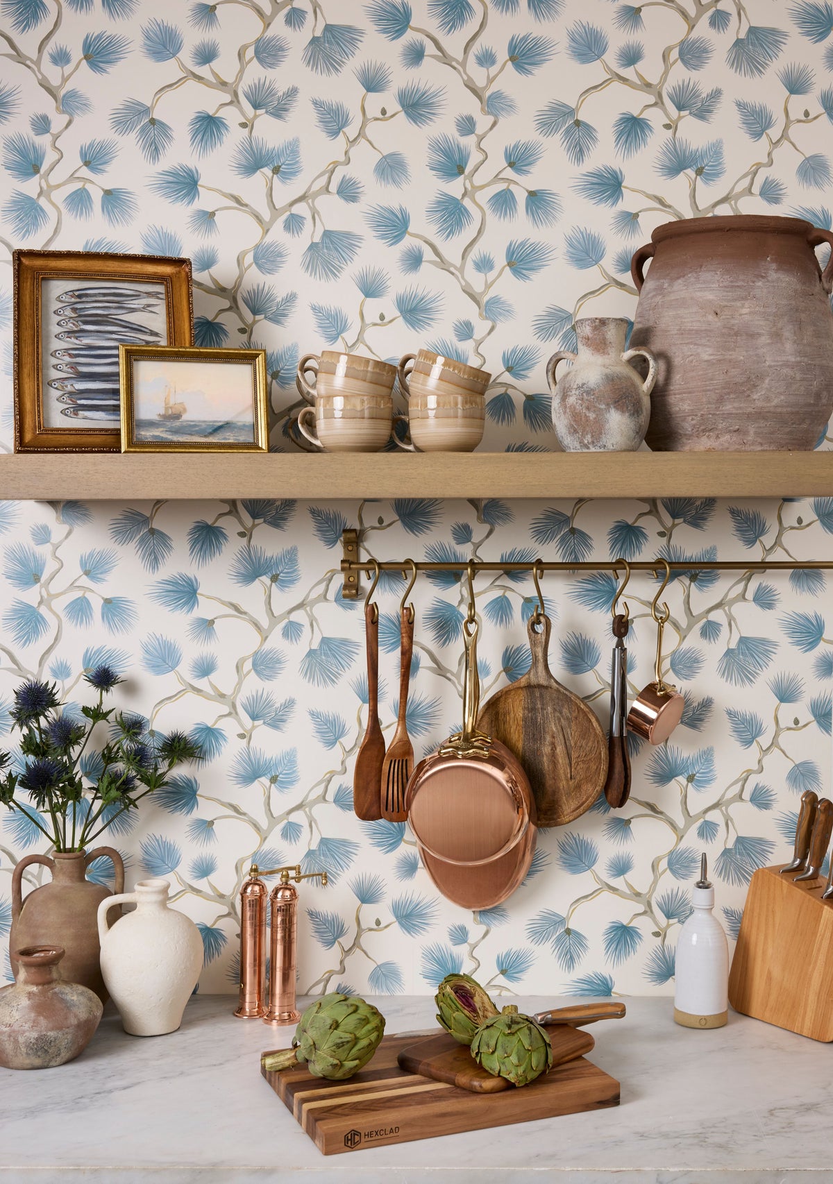 Rustic kitchen vignette with blue floral wallpaper, copper pots and mugs on shelves, wooden spoons and cutting board.