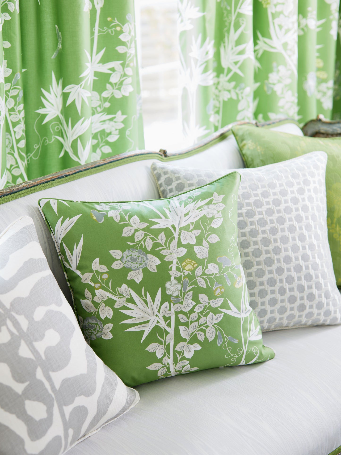 Green botanical print square cushion with white leaves on a light gray patterned sofa beside matching green curtains.