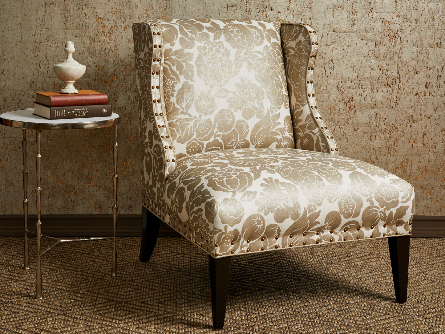 Beige damask upholstered wingback chair with nailhead trim and dark tapered legs beside a round marble-top side table with books and a white vase.