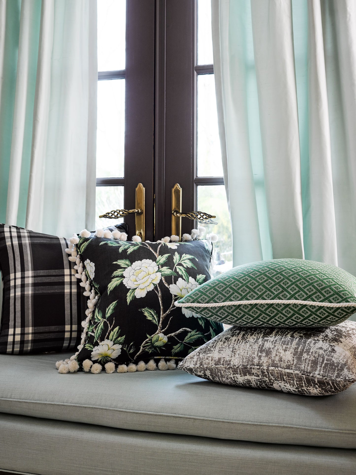 Mint curtains frame a window nook with floral pom-pom pillow, green geometric pillow, plaid pillow, and gray velvet cushion.