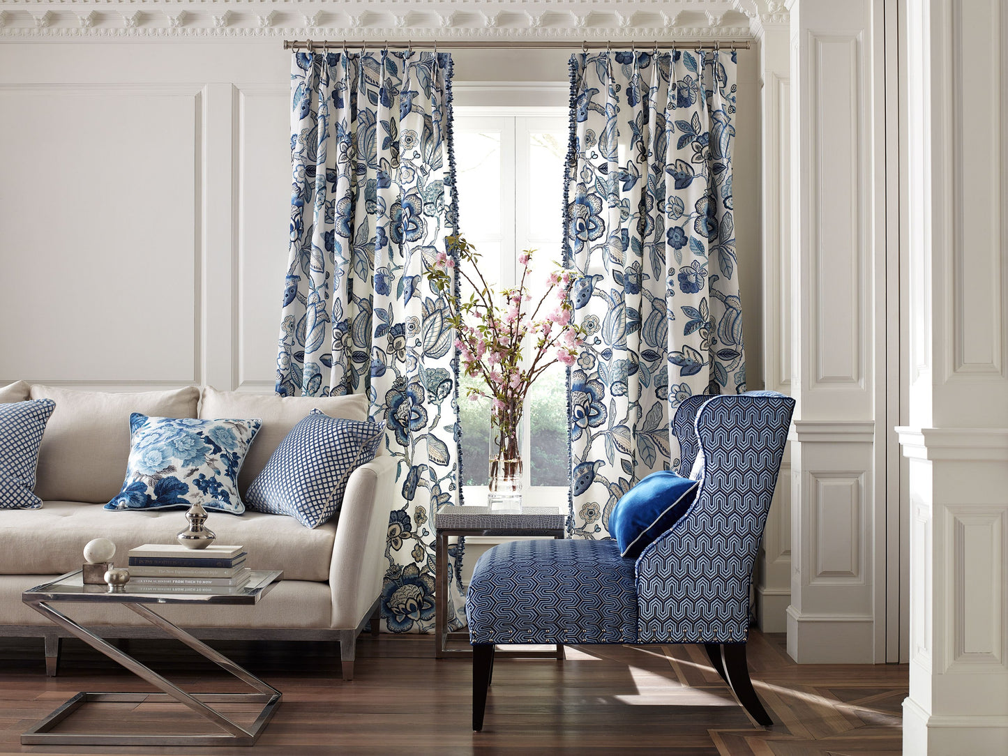 Living room scene with blue floral curtains, blue patterned accent chair, beige sofa, glass coffee table, vase with blossoms.