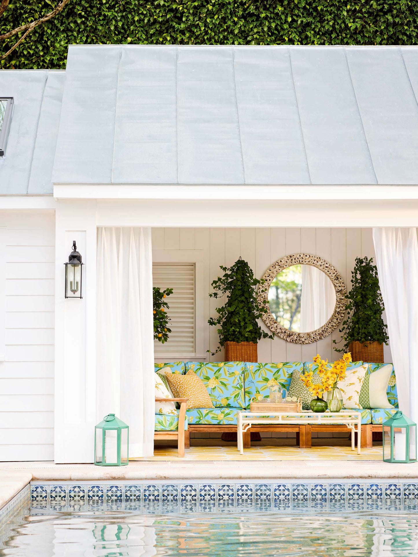 Poolside cabana with white curtains, tropical blue-green sofa, round wicker mirror, potted topiaries, turquoise lanterns.