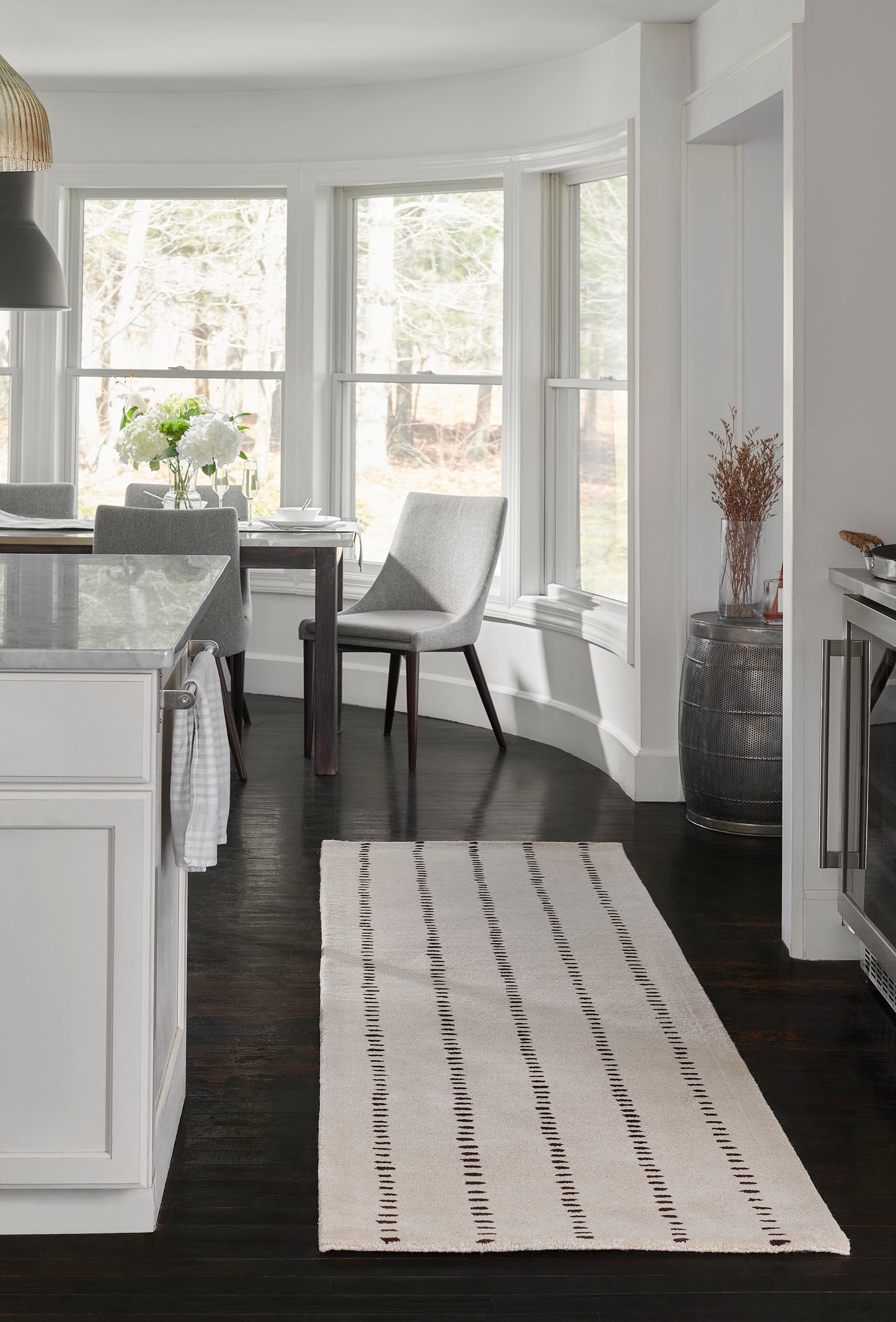 White rug with black stitched stripes on dark wood floor, near a marble island and grey dining chairs by bay windows.