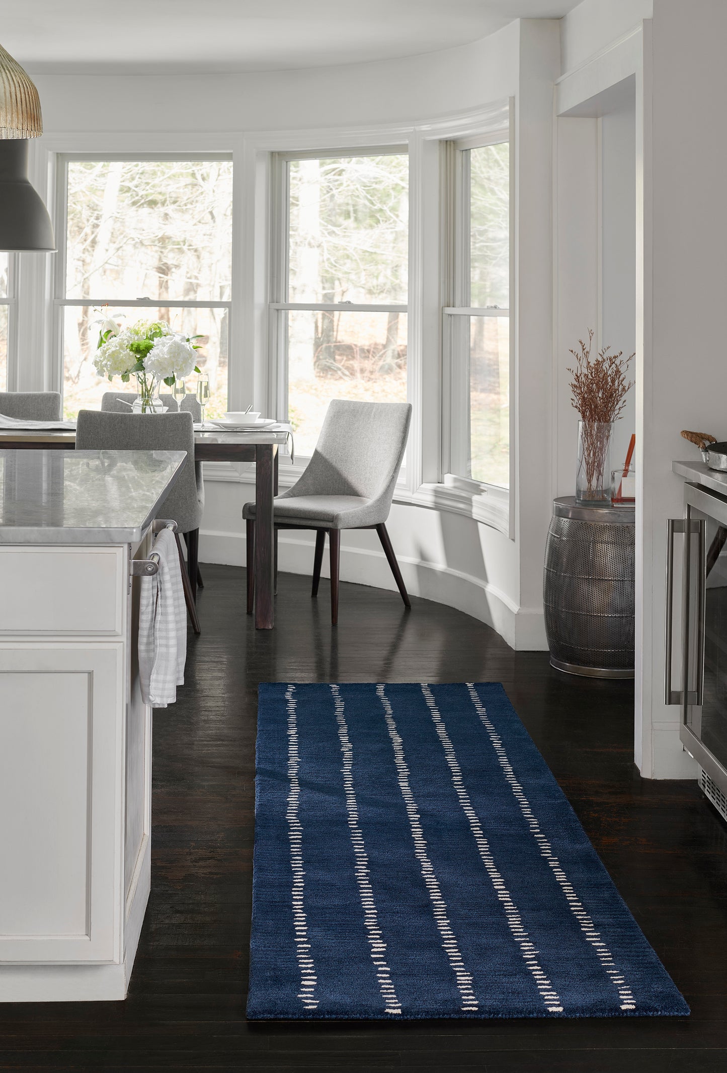 Blue striped rug on dark wood floor in a bright white kitchen; curved bay windows, gray chairs, and a marble island.