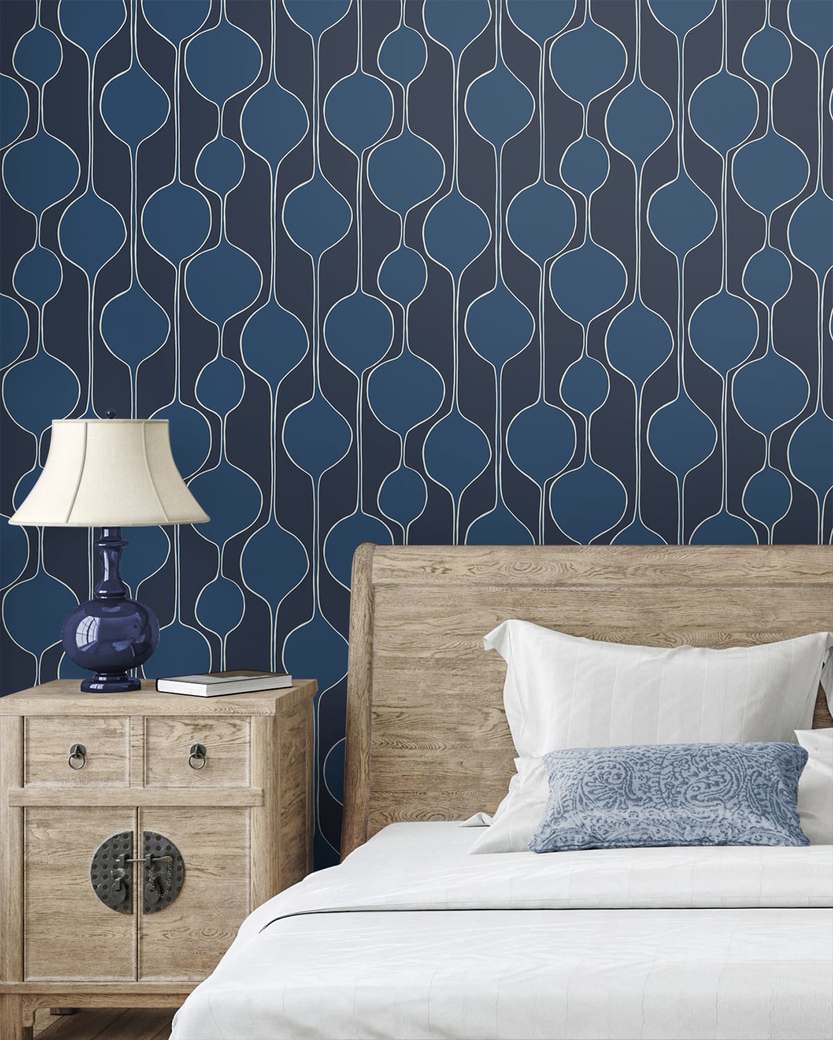 Bedroom with navy geometric wallpaper, light wood bed and nightstand, blue ceramic lamp, white bedding and blue patterned pillow.