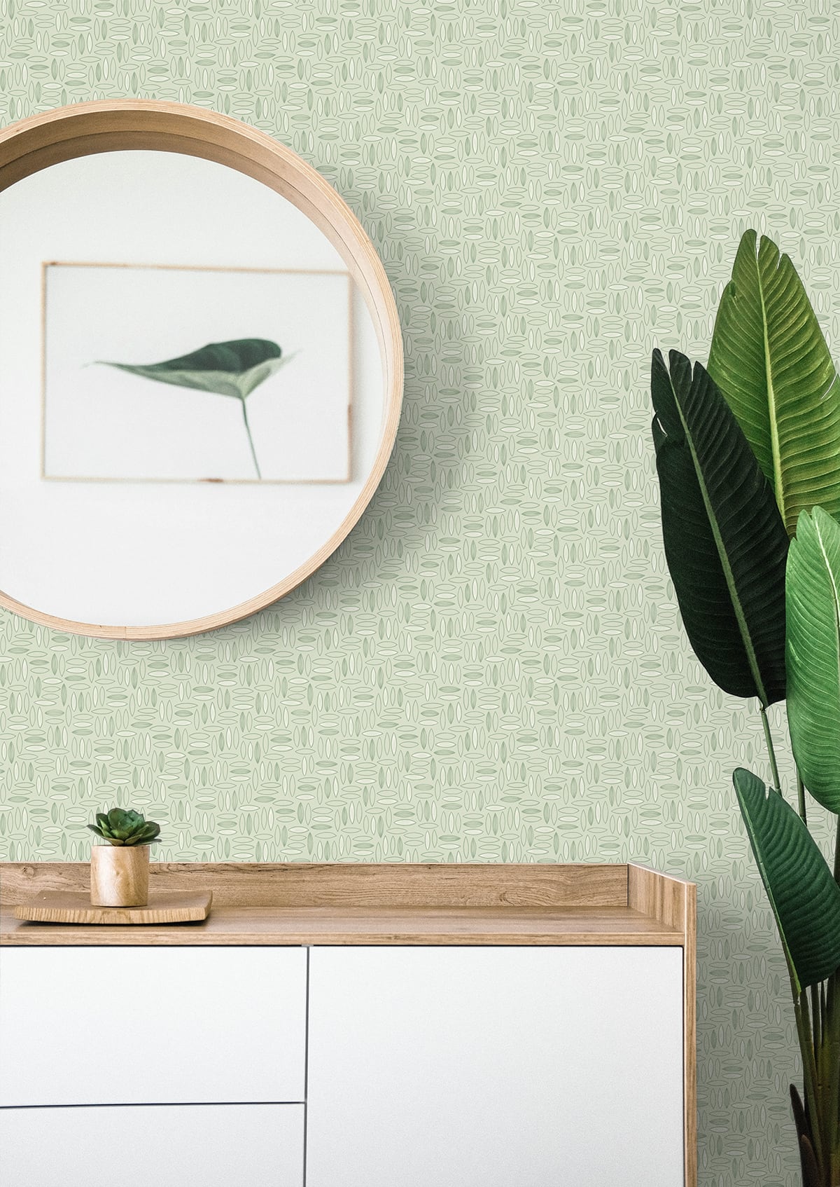 Geometric green matte wallpaper backdrop behind a round wooden mirror and a light wood console with white drawers; small succulent and tall leafy plant.