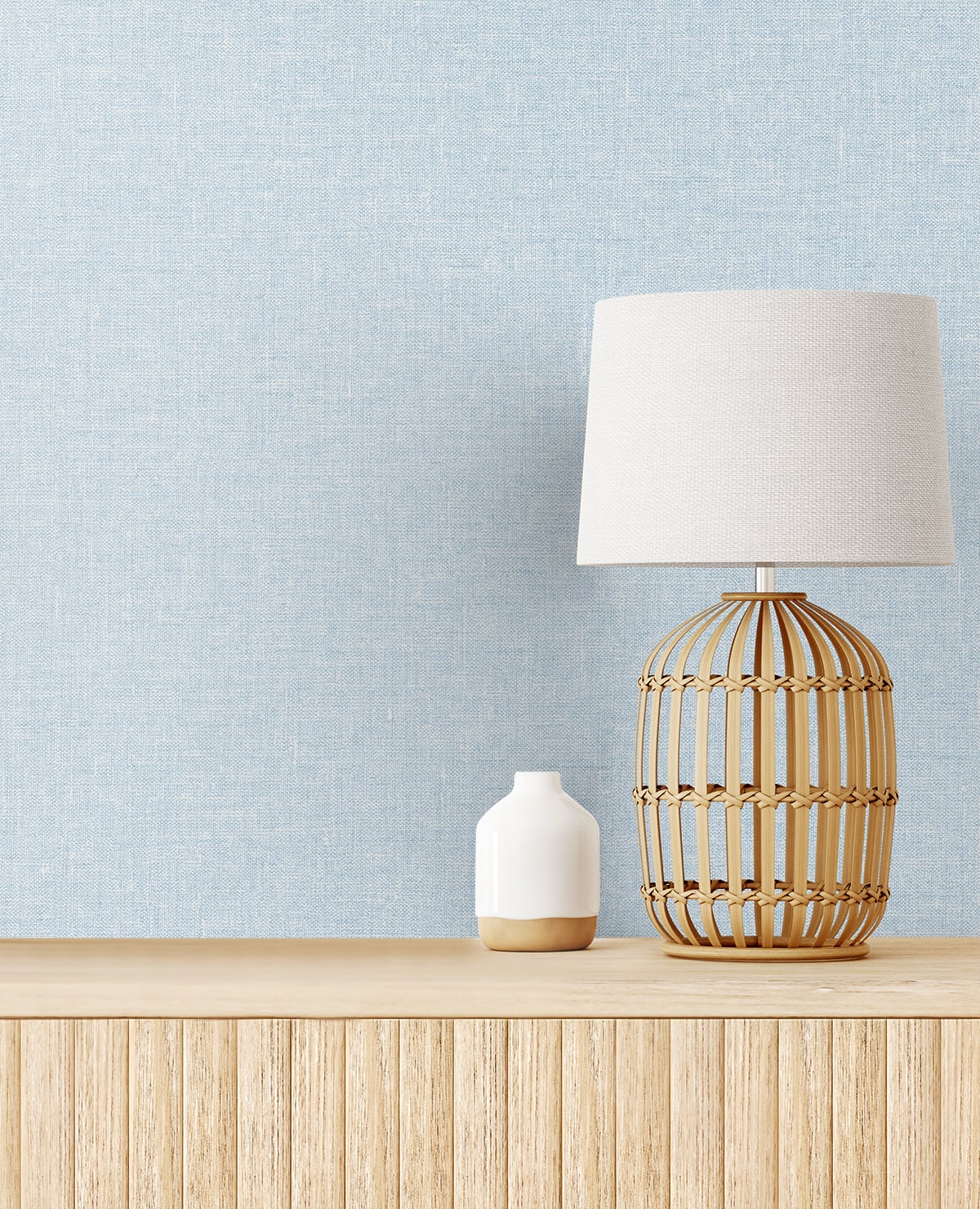 Soft blue matte linen textile product shown in a room setting with a boho rattan table lamp on a light wood sideboard, a small white ceramic vase, and a blue textured wall.
