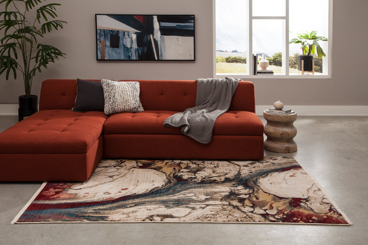 Red-orange tufted sectional with chaise, gray knit throw and patterned pillow; abstract wall art, round wooden side table, and multicolor area rug.