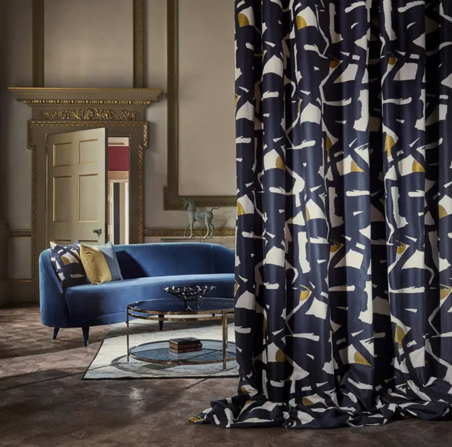 Navy blue abstract-pattern fabric drapery with white and gold accents beside a curved blue velvet sofa in a luxe living room