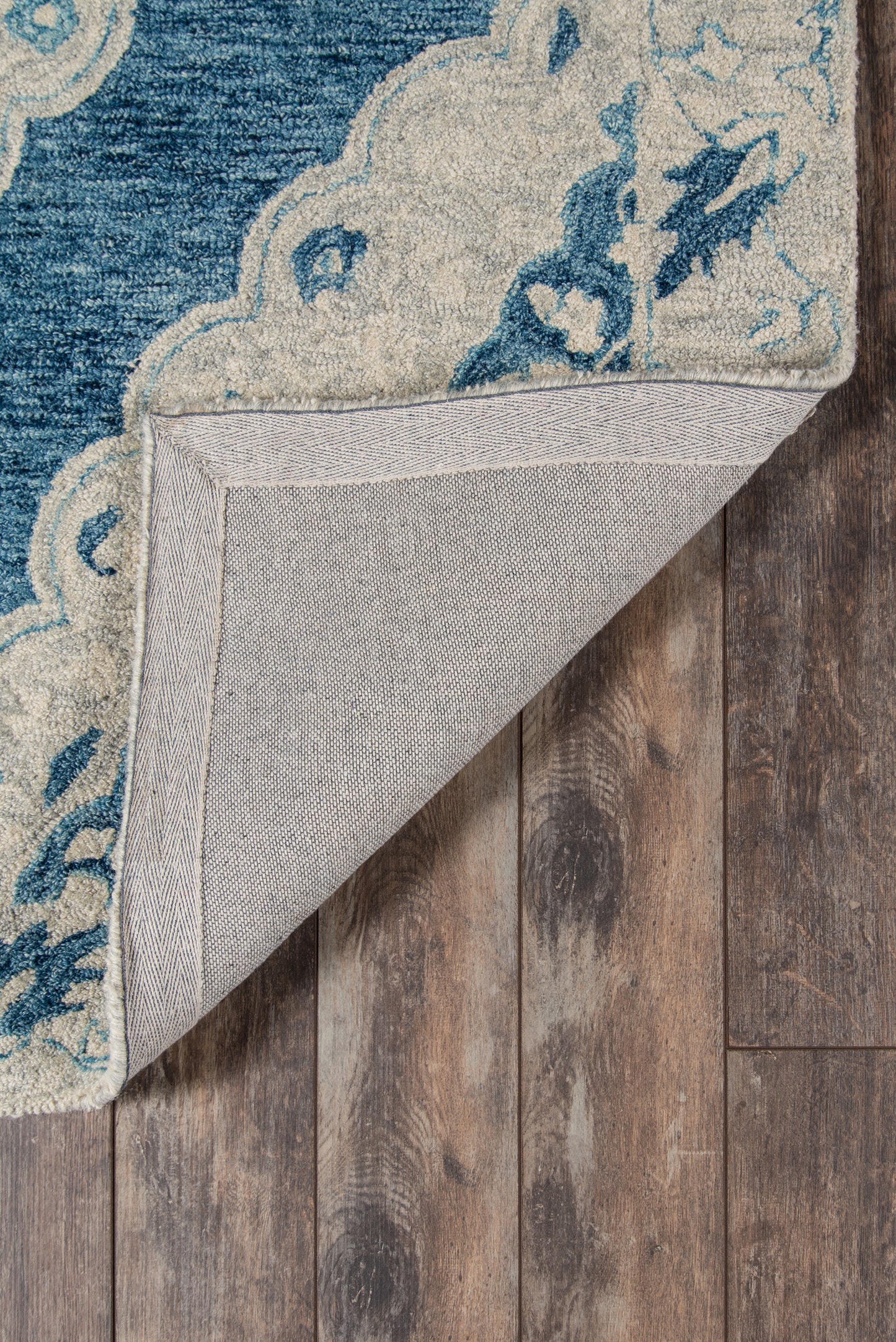 Blue and cream ornate rug with floral motifs; folded corner reveals light gray backing and herringbone edge on wood floor.