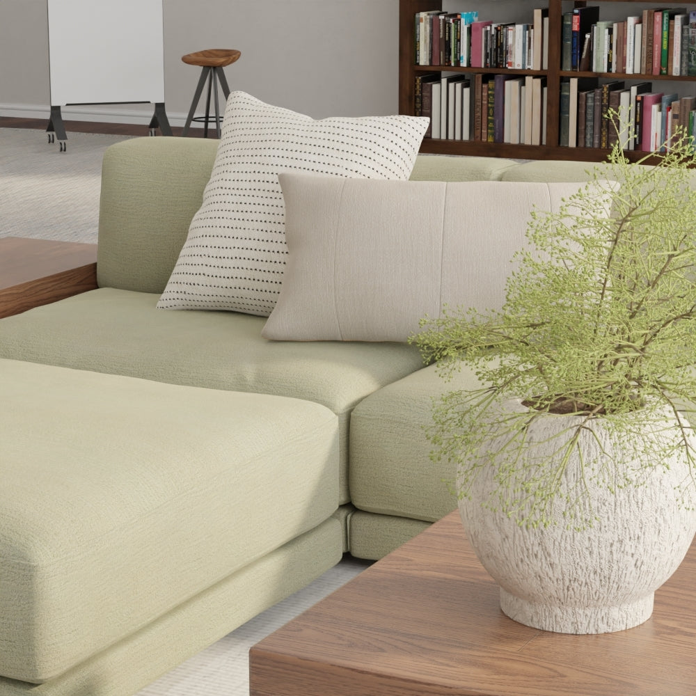 Sage solid fabric sofa with cream cushions and a dotted pillow; wooden side table with a white planter and greenery.