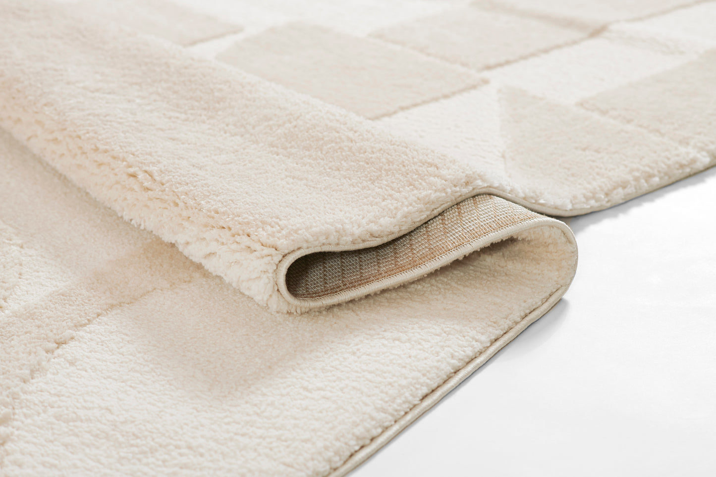 Beige plush area rug with a subtle square pattern; rolled edge reveals woven backing.