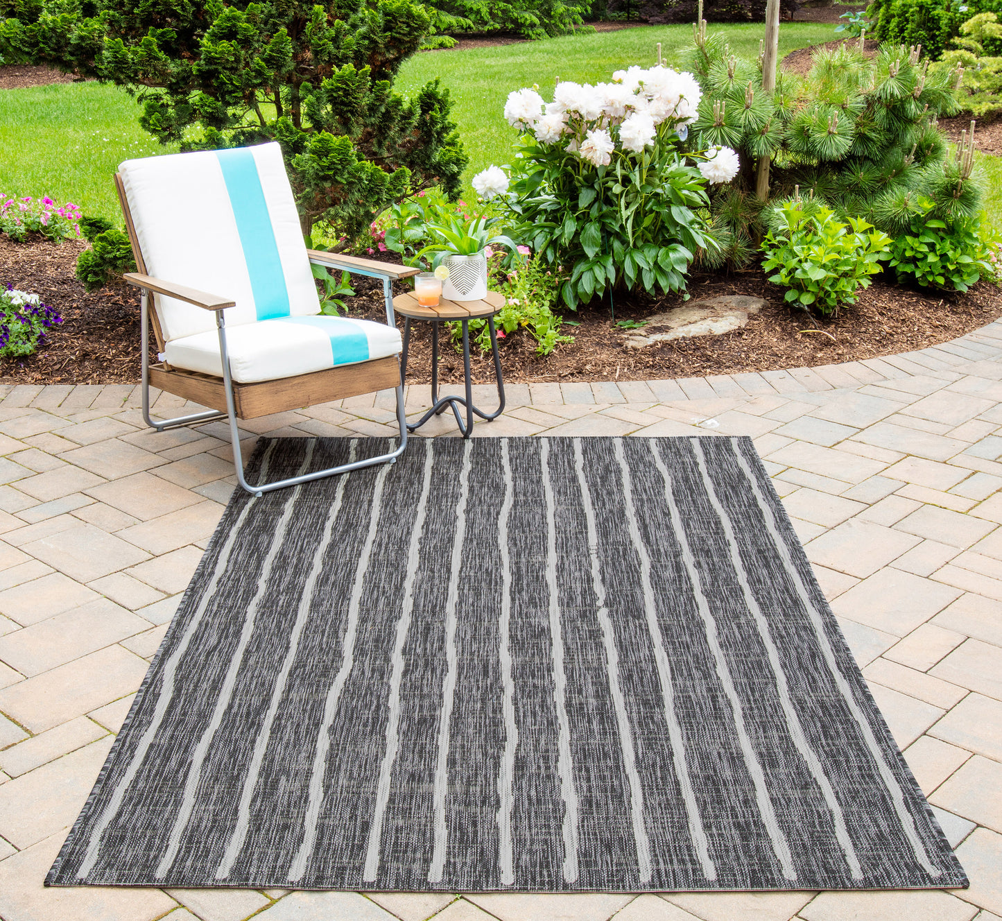 Outdoor grey striped rug on a paver patio beside a metal-framed chair with white cushions and blue stripe, garden backdrop.