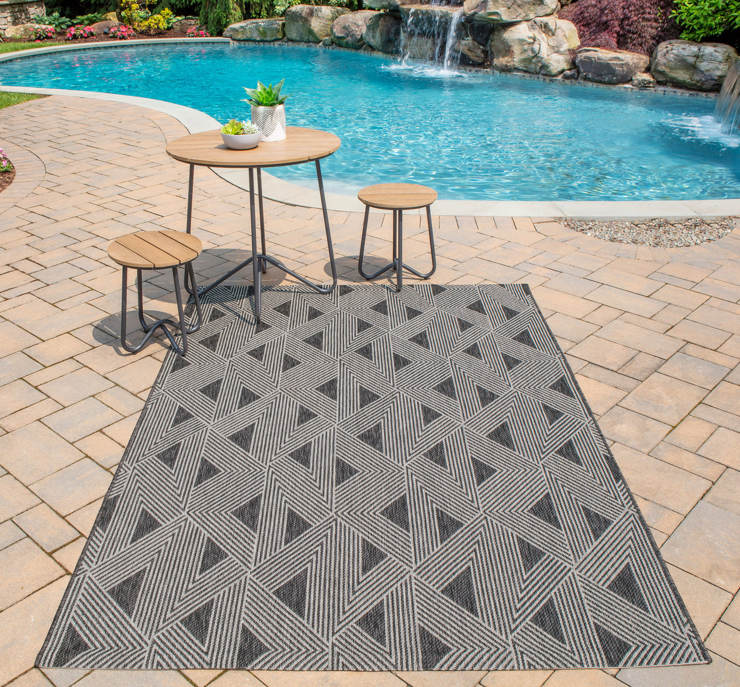 Outdoor rug with gray geometric triangle pattern on brick-paver patio, with round wood-top table and stools beside a pool.