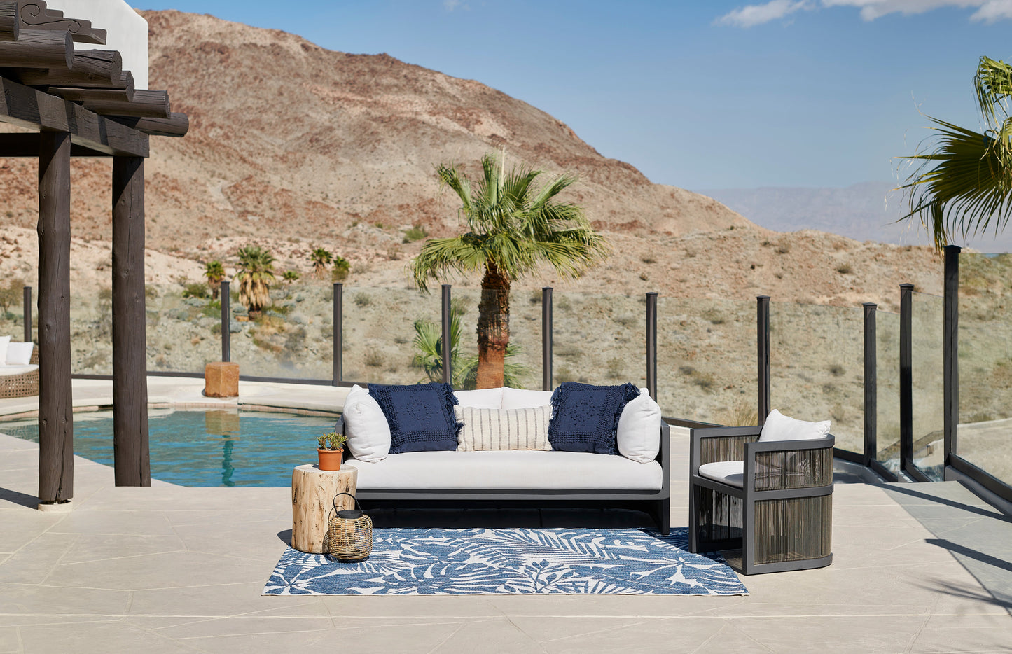 Blue botanical rug in a poolside outdoor lounge setting with a white cushioned sofa, navy pillows, dark side table, and palm trees.